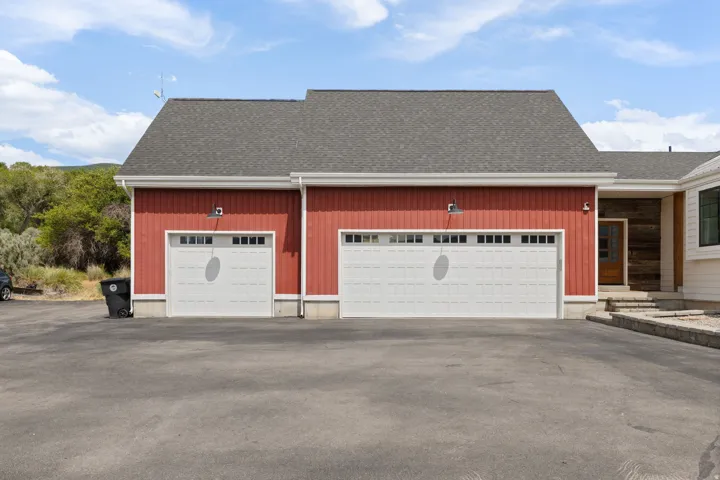 Exterior featuring two white garage doors with window inserts, red vertical siding, and a dark shingle roof