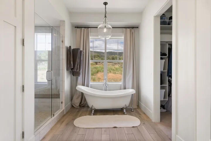 Freestanding clawfoot tub with a window backdrop, glass-enclosed shower, wood-finish flooring, and a built-in closet with shelving