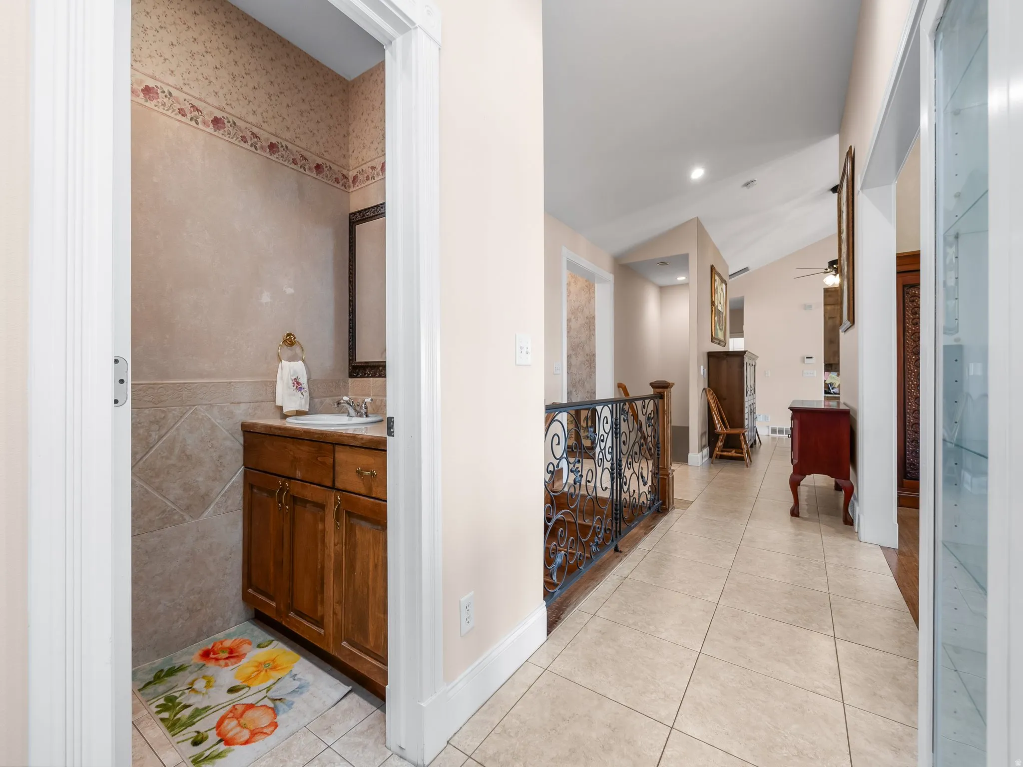 Hallway with neutral tile flooring leading to a powder room featuring a wood-finish vanity with a drop-in sink, a mirror with a decorative frame, and tiled walls with a patterned border