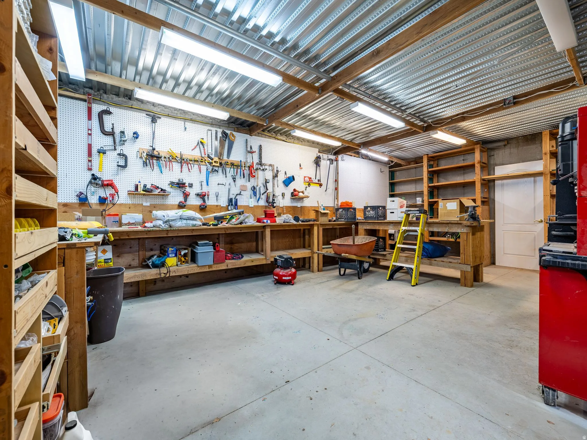Concrete-floored workshop featuring extensive built-in shelving, a large workbench with pegboard organization, fluorescent overhead lighting, and a corrugated metal ceiling