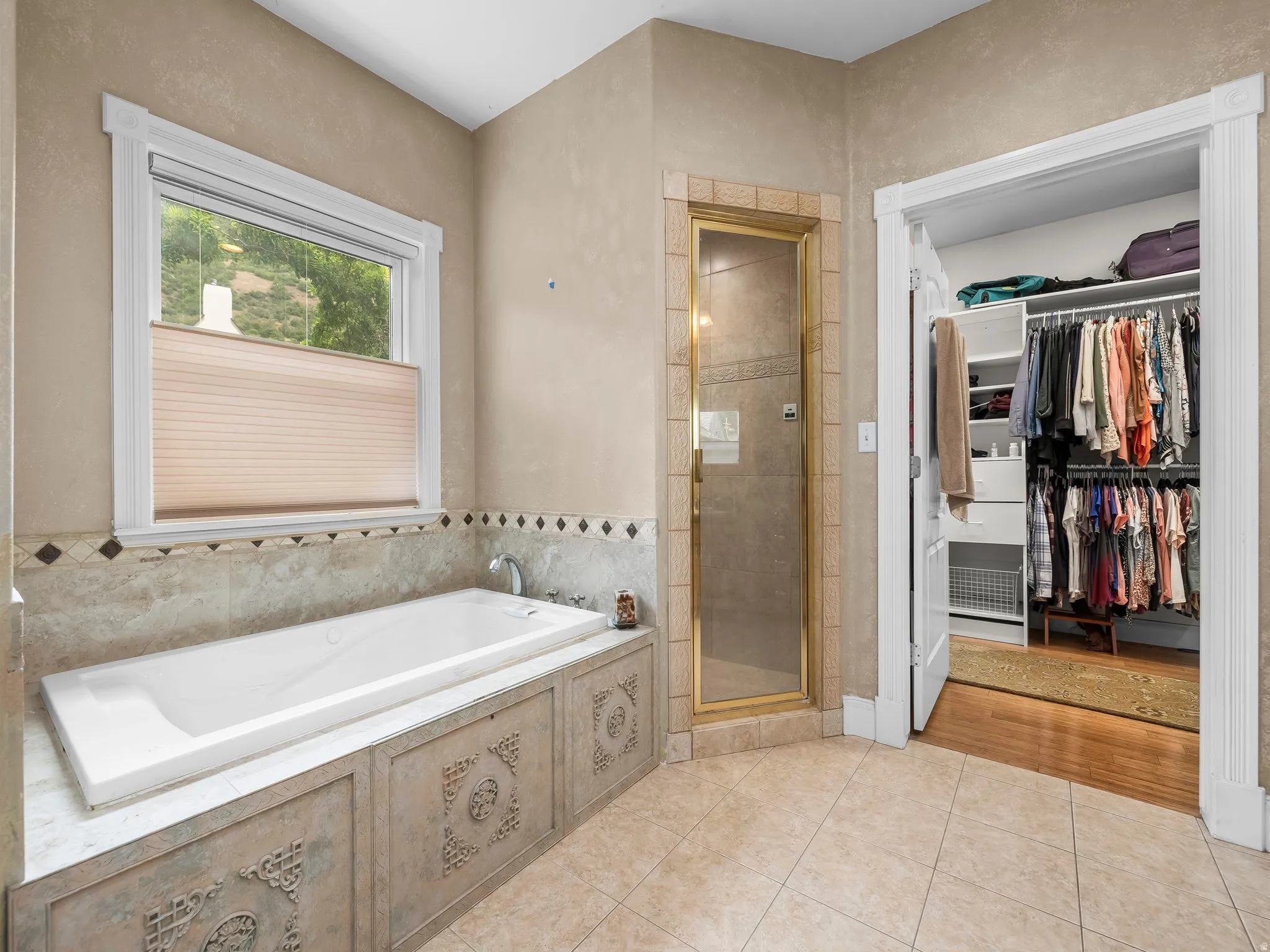 Spacious bathroom featuring a built-in soaking tub with decorative tile surround, a glass-enclosed shower with gold-tone trim, and neutral tile flooring