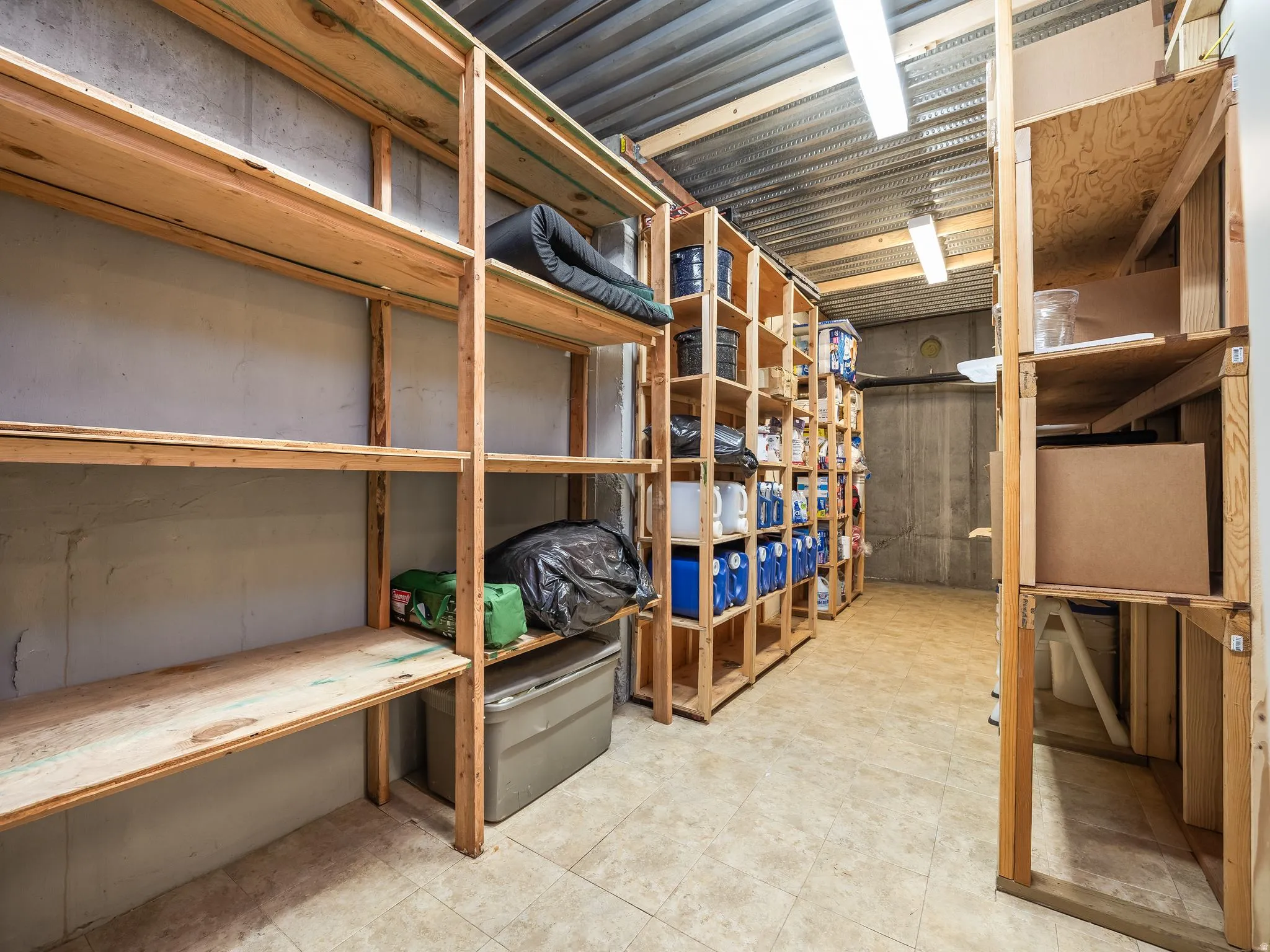 Dedicated storage room featuring extensive wood shelving, tiled flooring, exposed concrete walls, and overhead fluorescent lighting