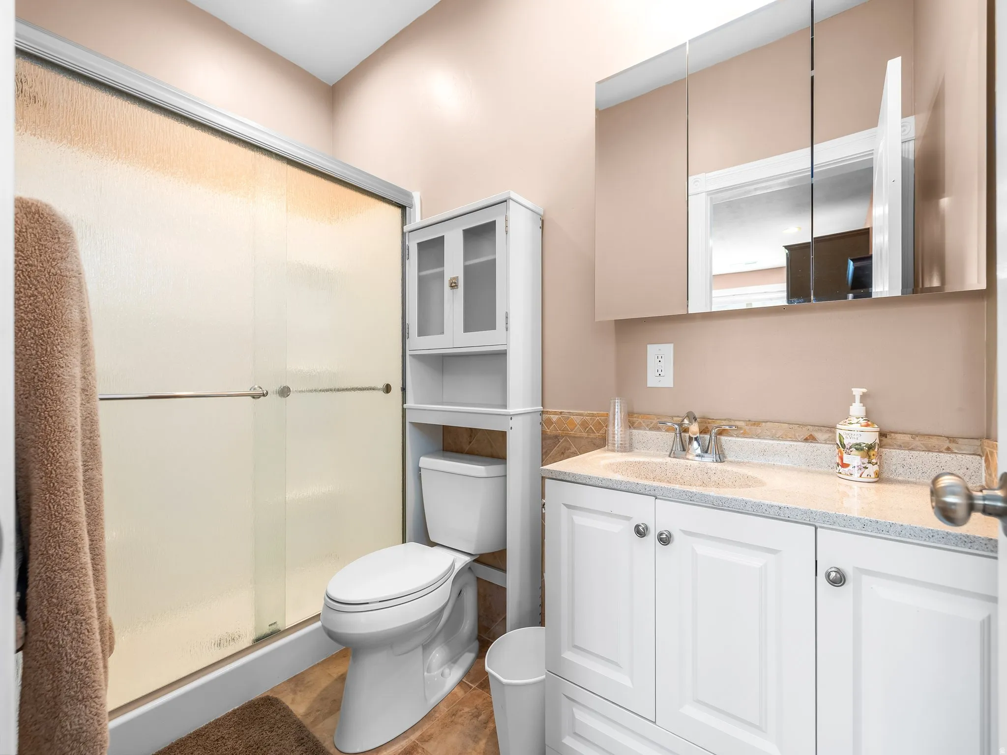 Bathroom featuring a white vanity with a speckled countertop, a mirrored medicine cabinet, a shower with translucent sliding doors, and a freestanding white storage cabinet
