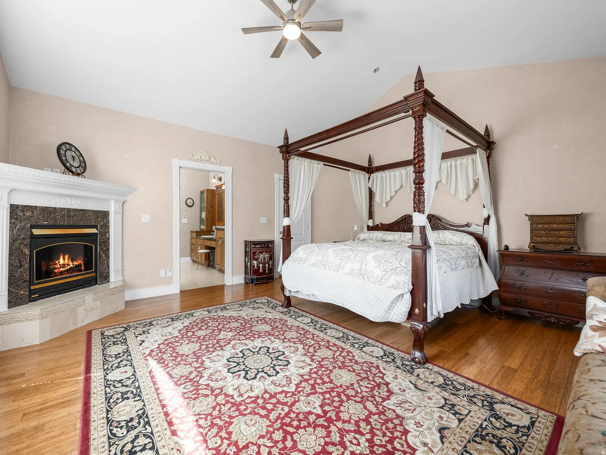 Primary bedroom with vaulted ceiling, ceiling fan, fireplace with decorative mantel, wood-finish flooring, and an open doorway revealing primary bathroom with a vanity
