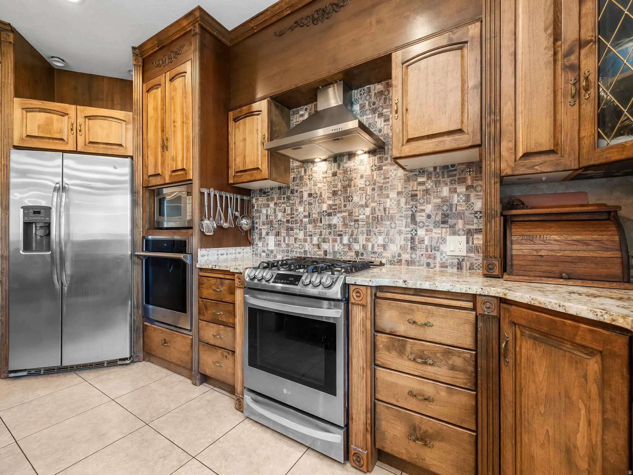 Kitchen featuring custom wood cabinetry, stainless steel appliances, patterned tile backsplash, granite countertops, and light-toned floor tiling