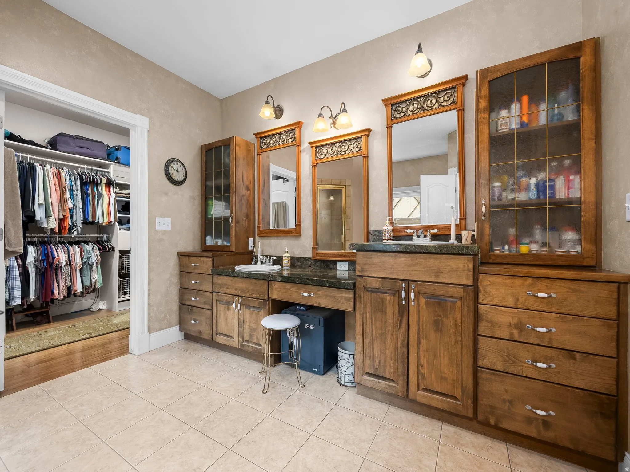 Primary bathroom featuring twin wood vanities with undermount sinks and dark countertops, individual mirrors, built-in cabinetry with glass doors, and light tile flooring