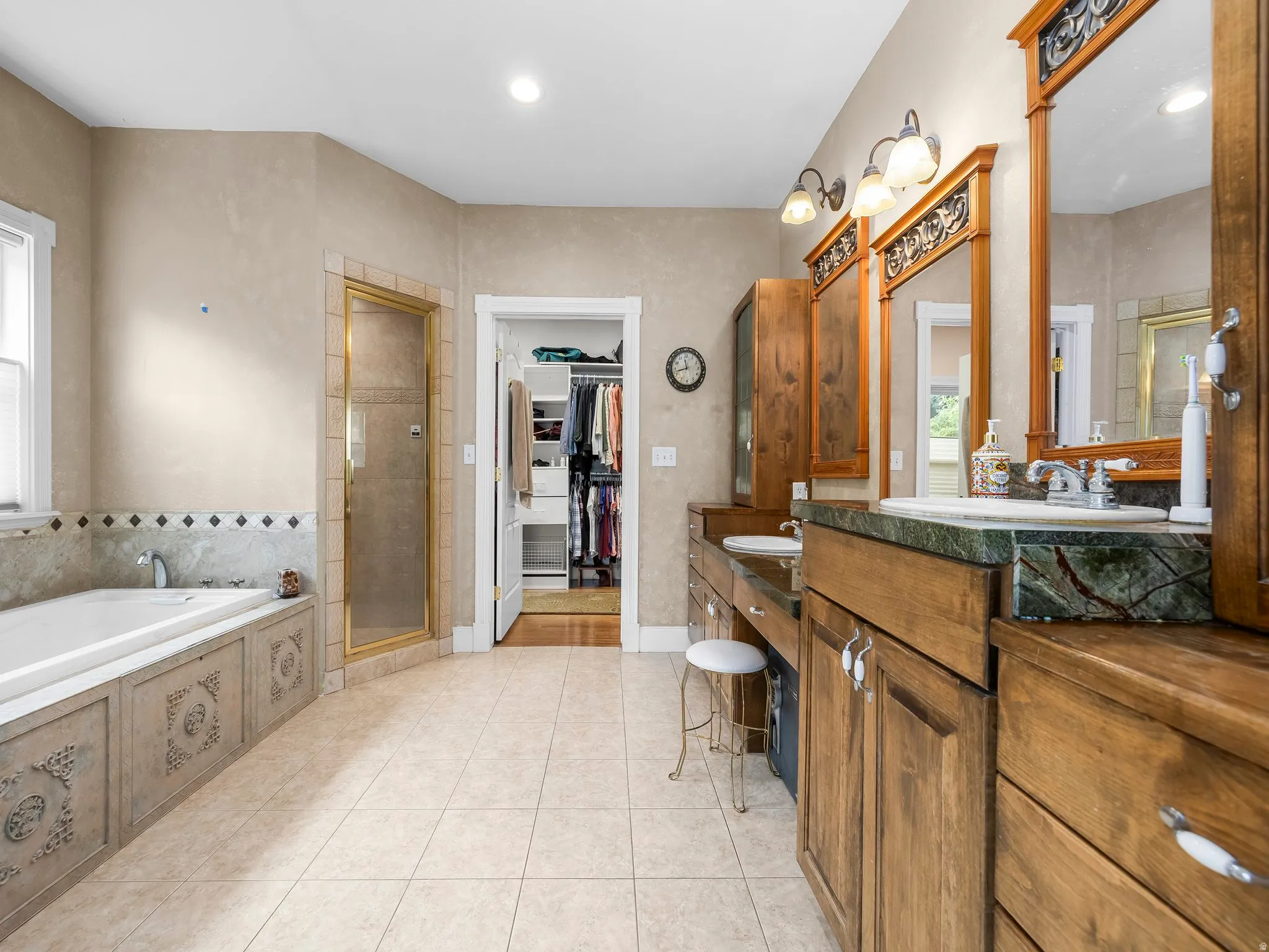 Primary bathroom featuring a dual vanity with green stone countertops, wood cabinetry, a built-in soaking tub with decorative paneling, a glass-enclosed shower, and tile flooring