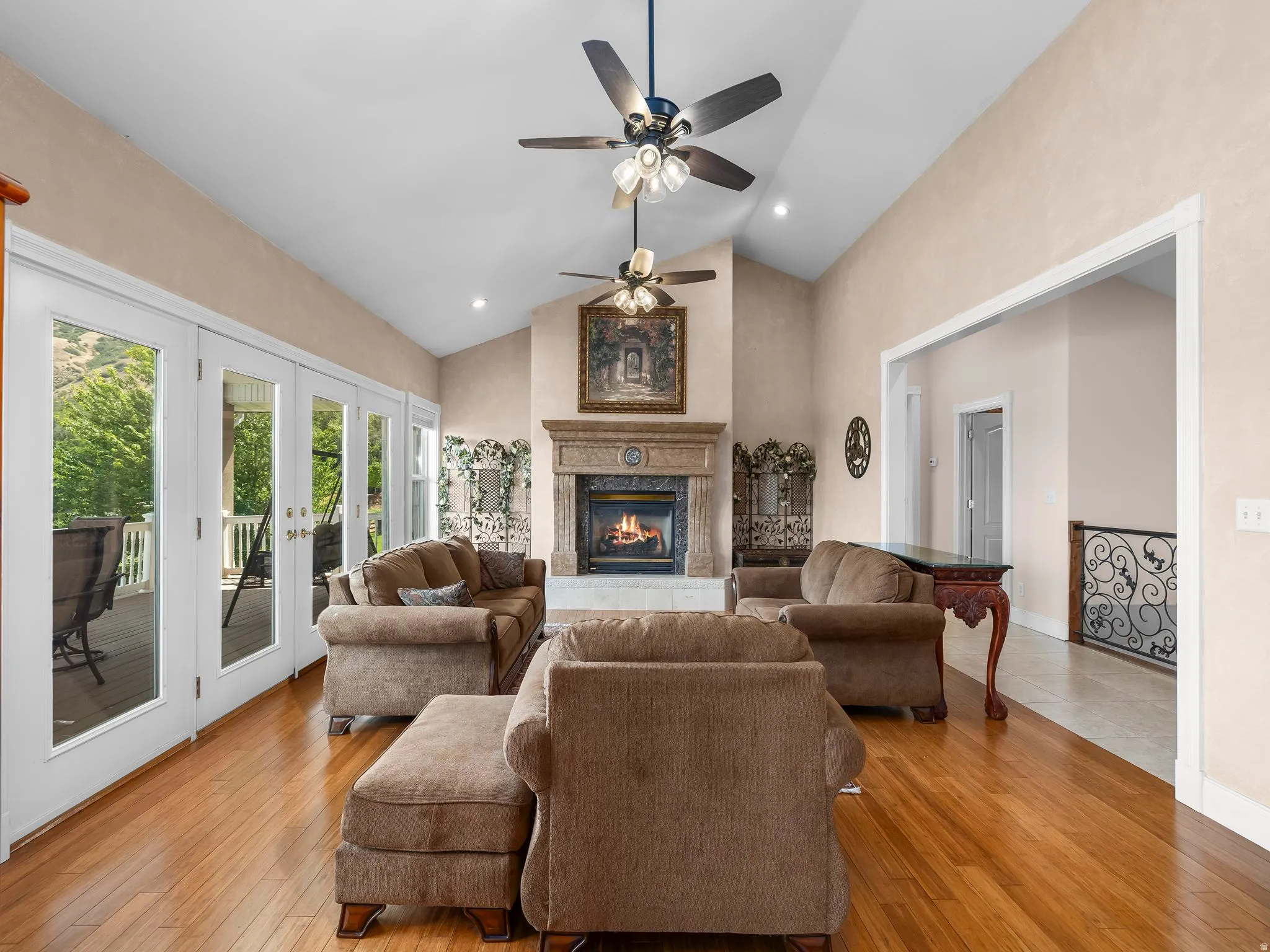 Living area featuring a vaulted ceiling with two ceiling fans, wood-finish flooring, a stone fireplace with ornate mantel, and multiple glass-paneled French doors opening to an outdoor deck
