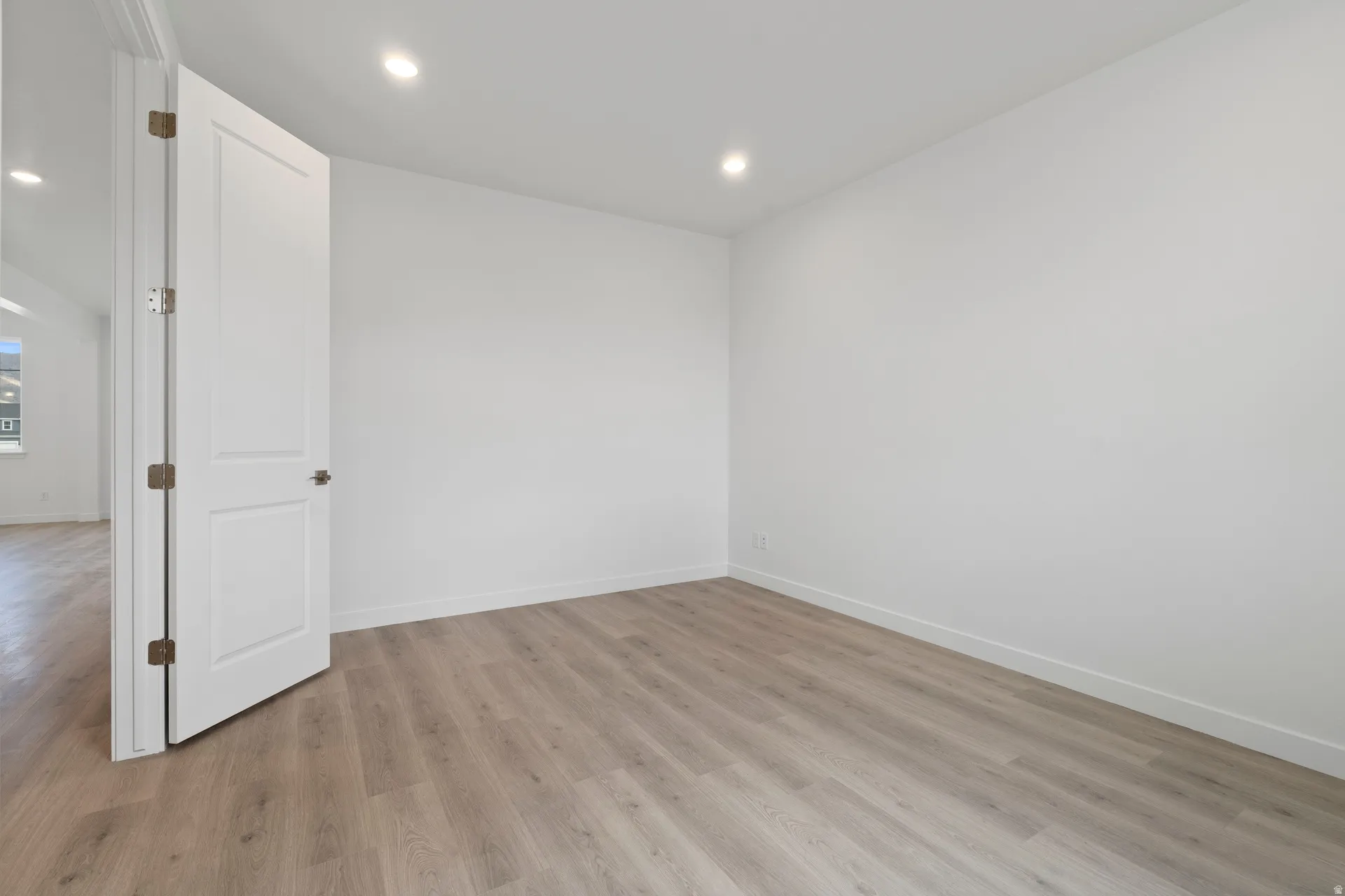 Room featuring light wood-finish flooring, white walls, recessed ceiling lighting, and a white two-panel interior door with bronze hardware