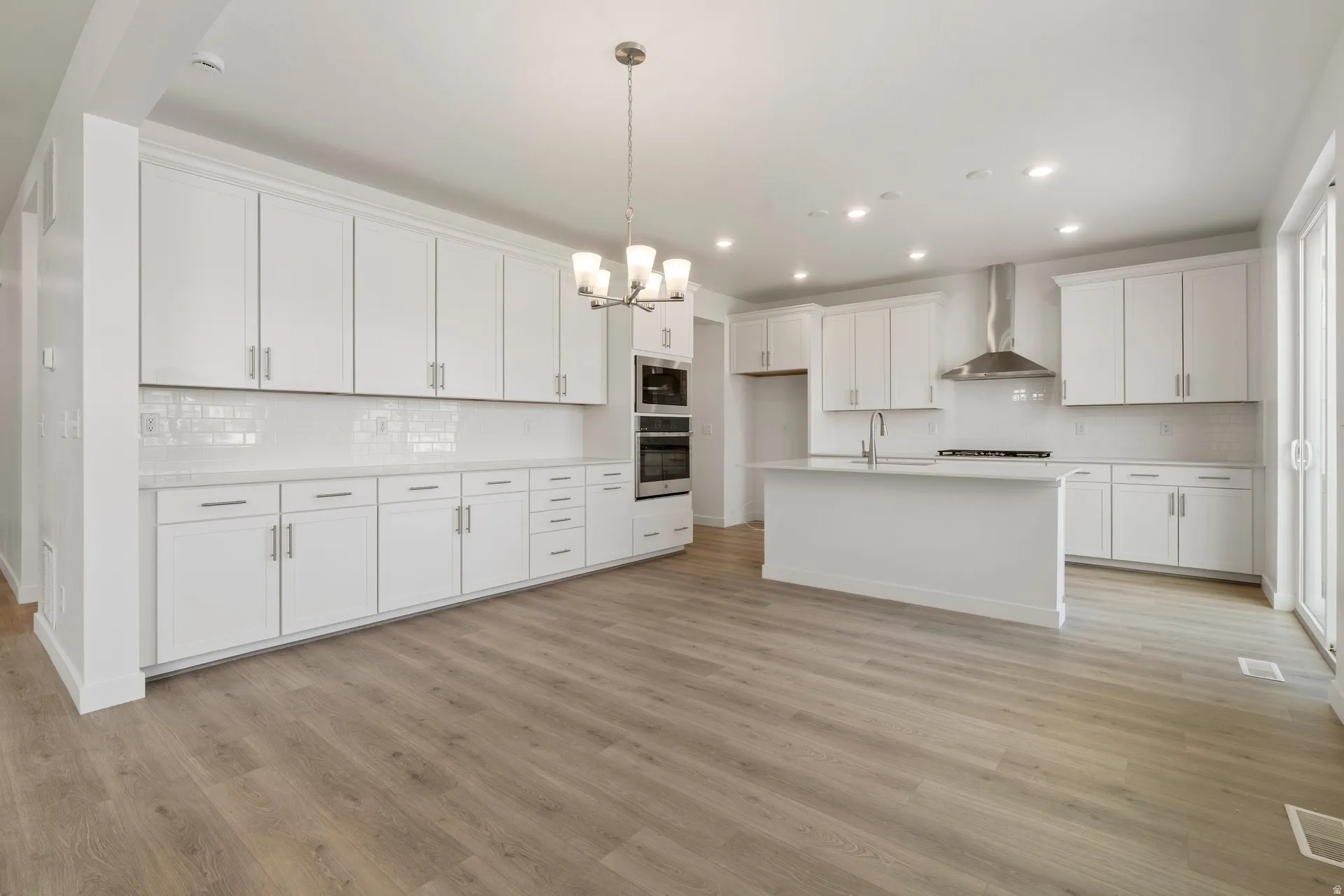 Spacious kitchen featuring extensive white cabinetry, light wood-finish flooring, a central island with sink, built-in double ovens, and a stainless steel range hood