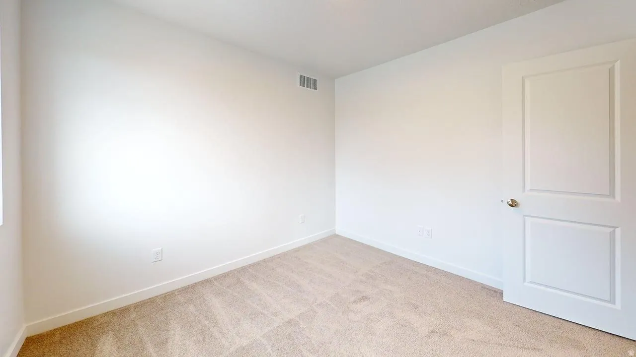 Room featuring neutral-toned walls, plush carpeting, a white paneled door with a brass-toned doorknob, white baseboards, and a visible air vent