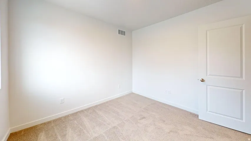 Room featuring neutral-toned walls, plush carpeting, a white paneled door with a brass-toned doorknob, white baseboards, and a visible air vent