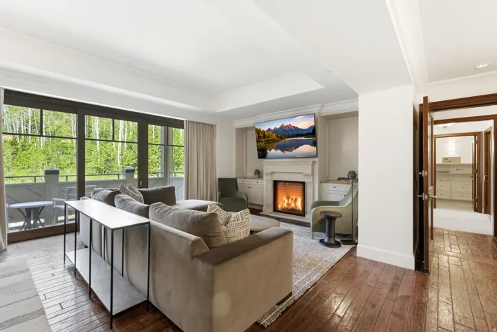 Living area with wood-finish flooring, a built-in fireplace with a white mantel, and extensive windows featuring a natural outdoor backdrop