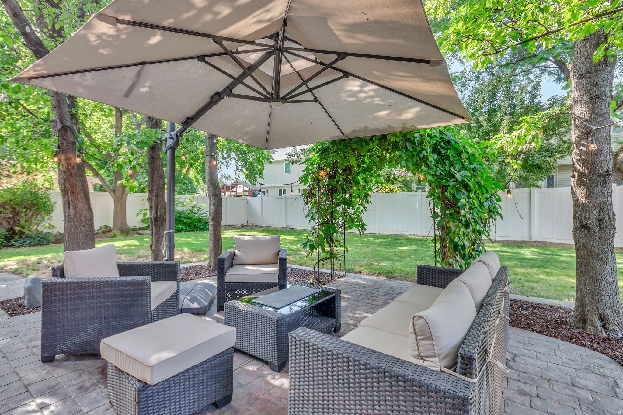 Paved patio area featuring a large cantilever umbrella, mature shade trees, a lush green lawn, and a white privacy fence