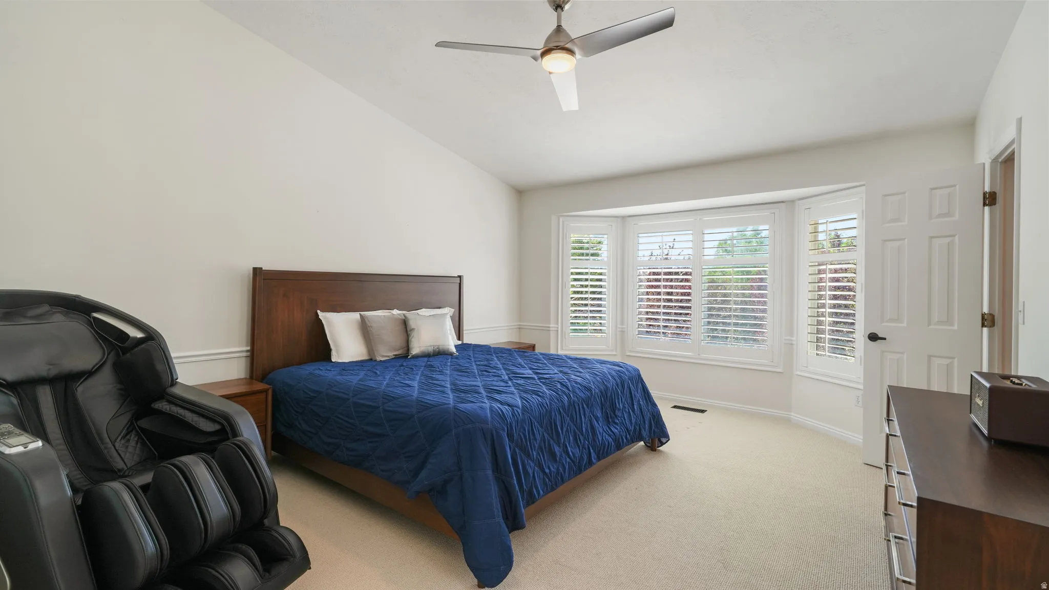Vaulted ceiling with a contemporary ceiling fan, bay window with plantation shutters, light-toned carpeting, and a paneled interior door
