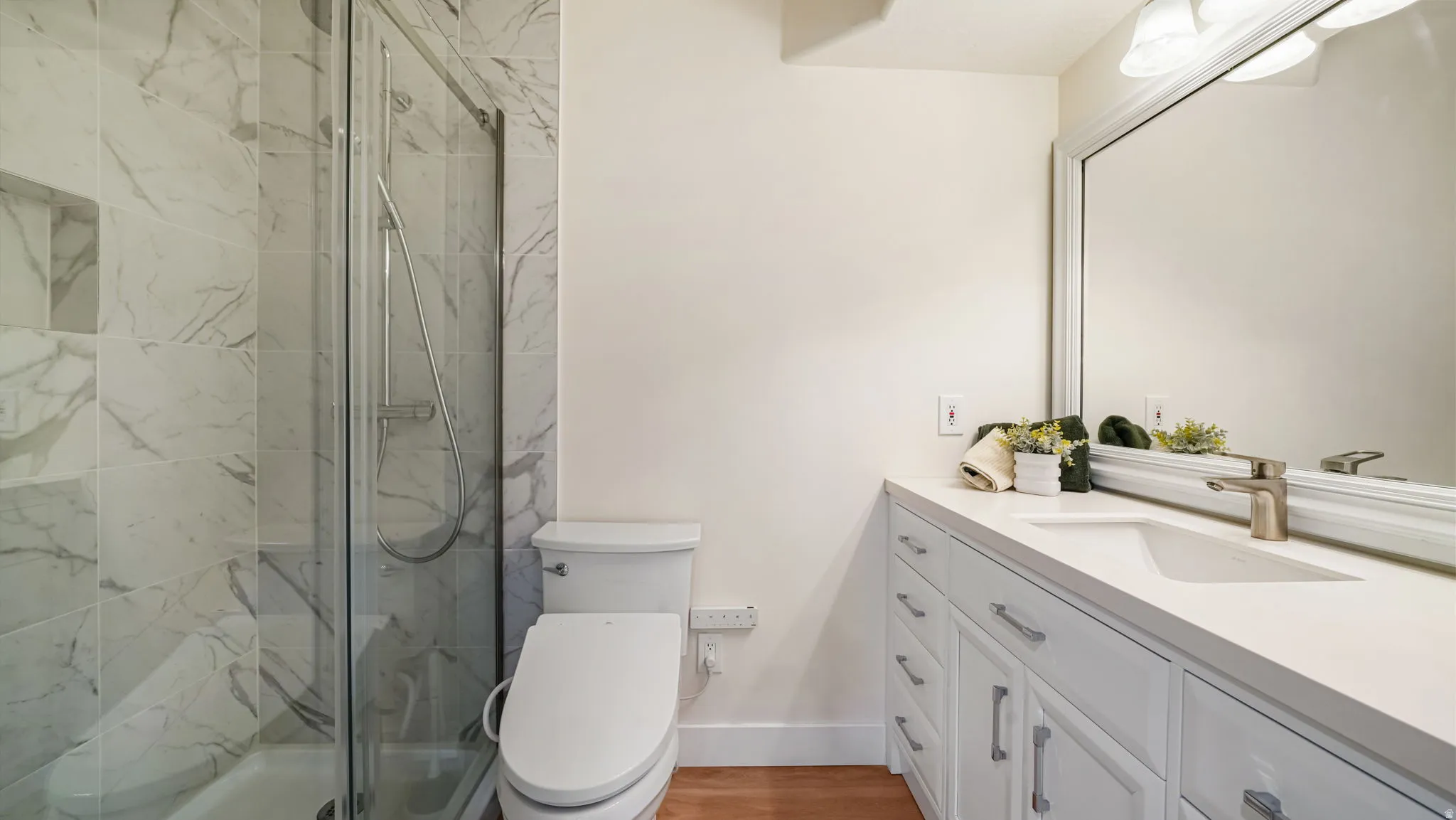Bathroom featuring a glass-enclosed shower with tiled walls and a recessed niche, a white vanity with an integrated sink, a large framed mirror, and wood-finish flooring