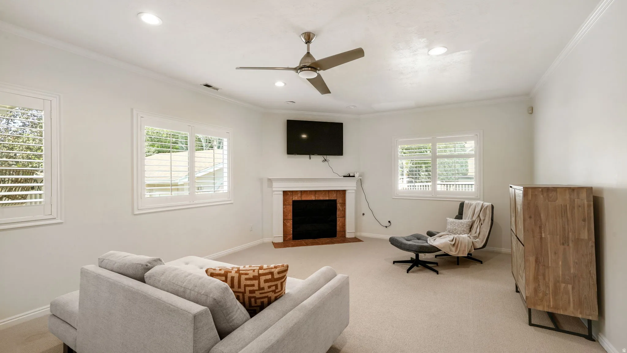 Spacious living area featuring a central fireplace with a brick surround and white mantel, plantation shutters on multiple windows, recessed lighting, and a ceiling fan