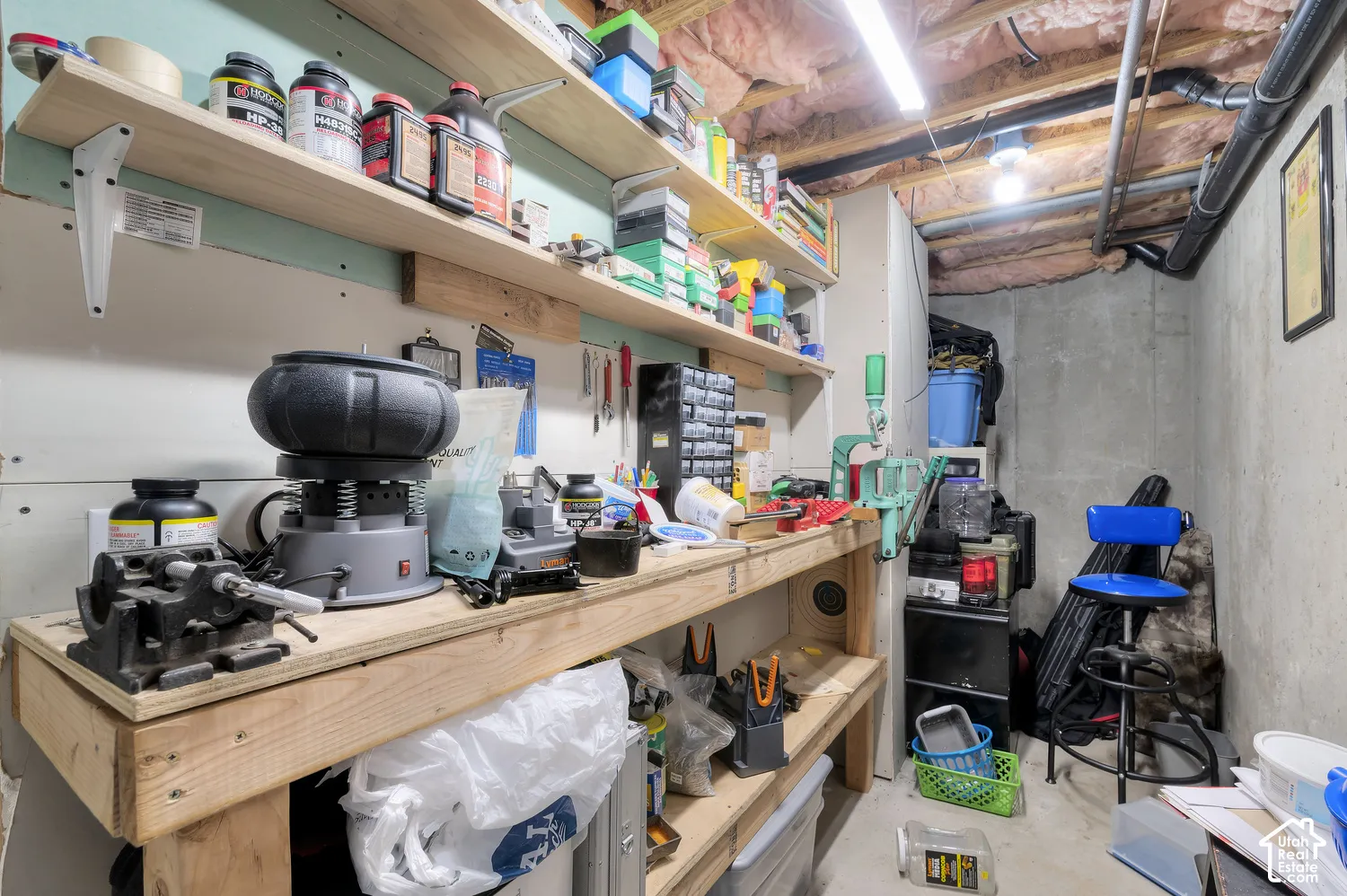 Basement workshop featuring a built-in wooden workbench, extensive shelving, concrete walls, exposed ceiling joists, and overhead lighting
