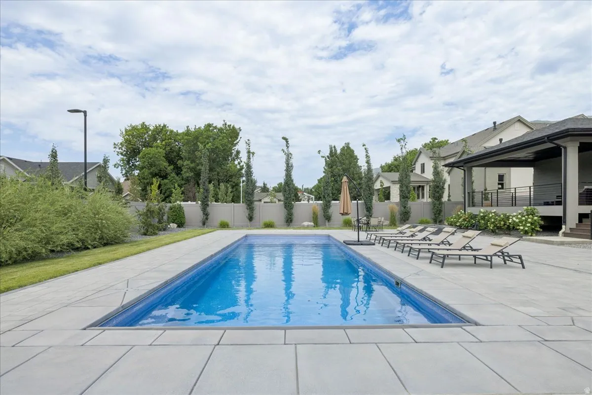 Rectangular swimming pool with expansive light-toned paver surround, bordered by mature trees and a privacy fence