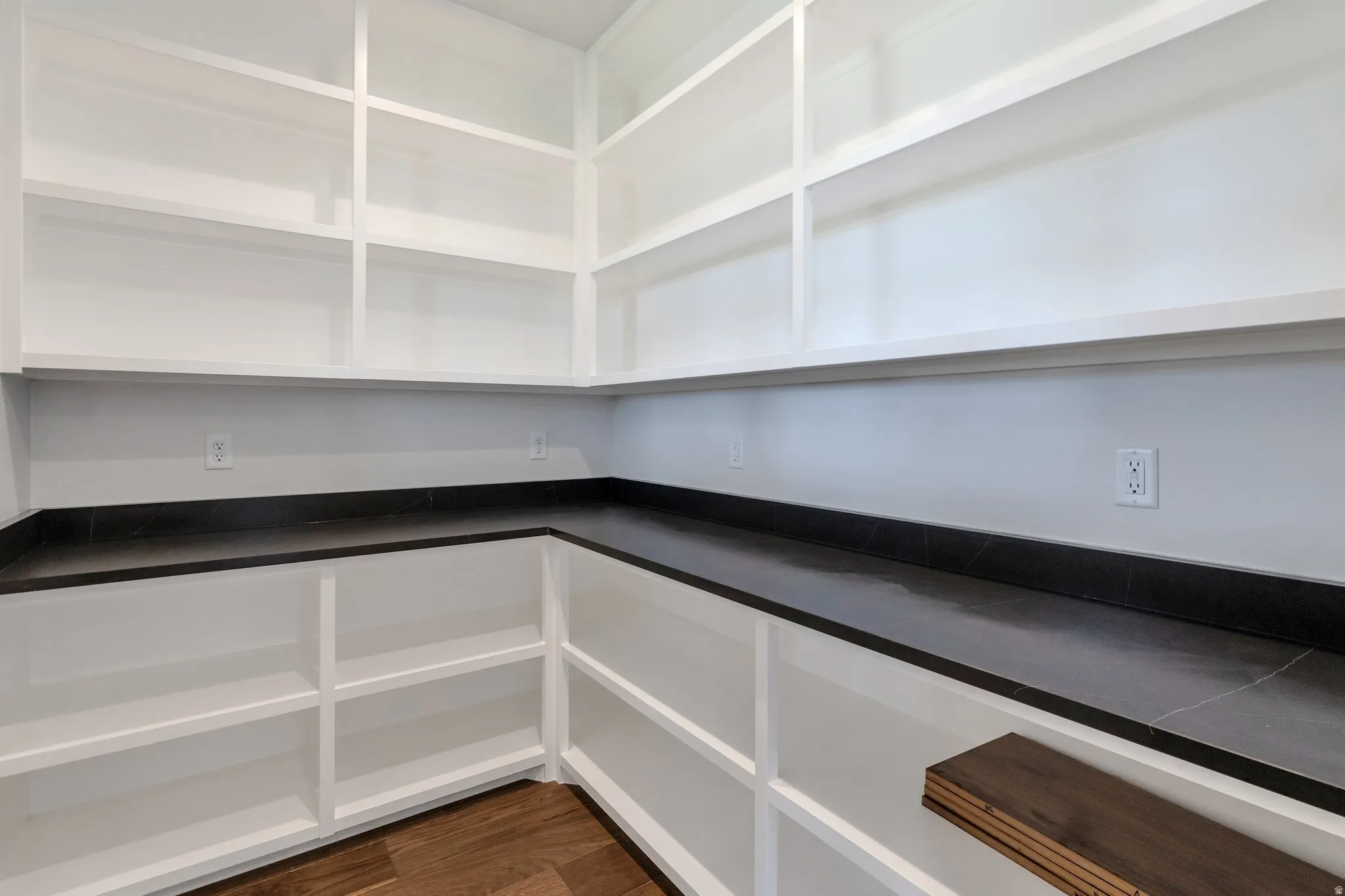 Expansive walk-in pantry featuring white built-in shelving, dark countertops, and wood-finish flooring