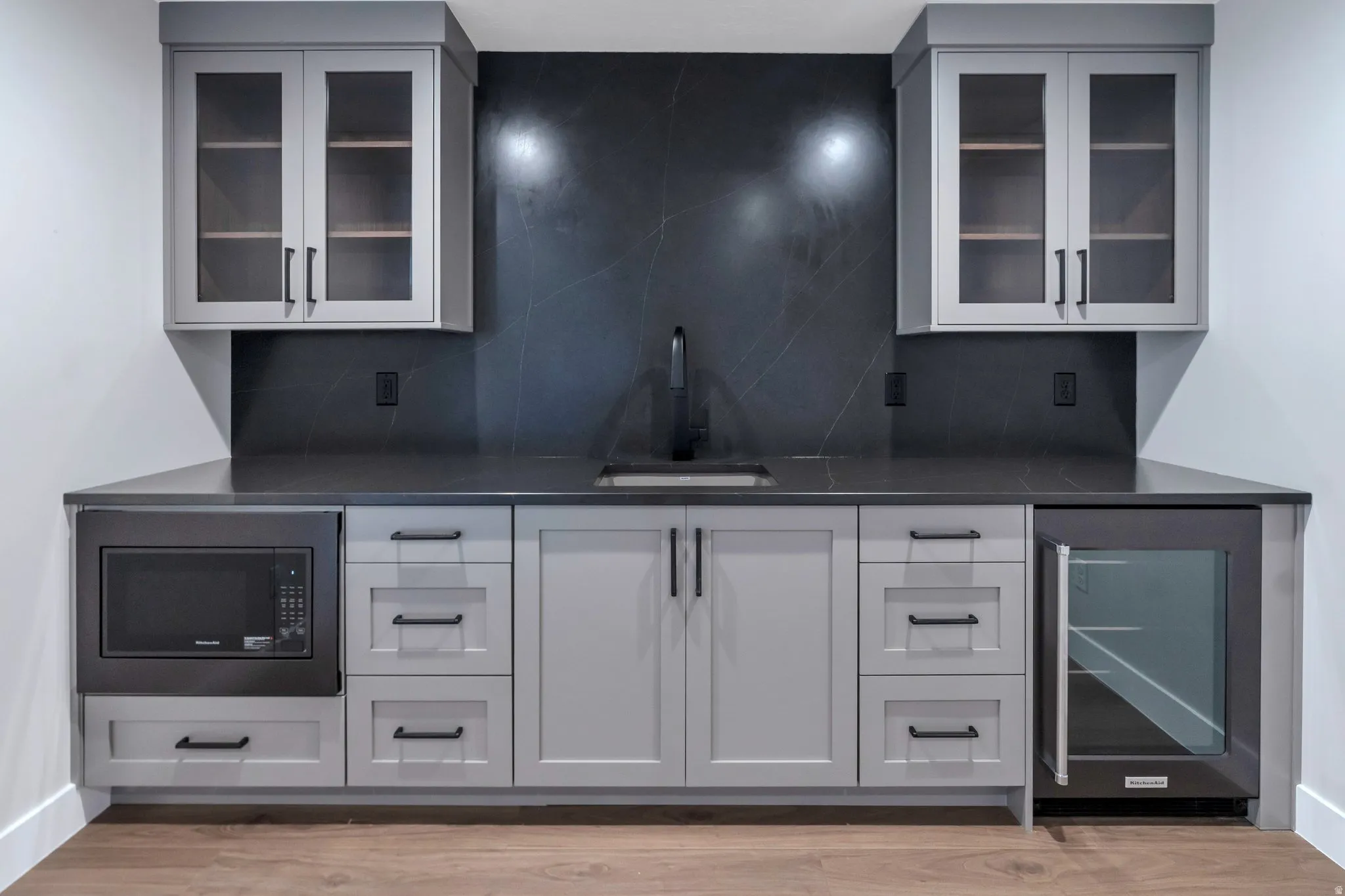 Contemporary wet bar featuring light gray cabinetry with dark hardware, dark countertop, and a dark tile backsplash