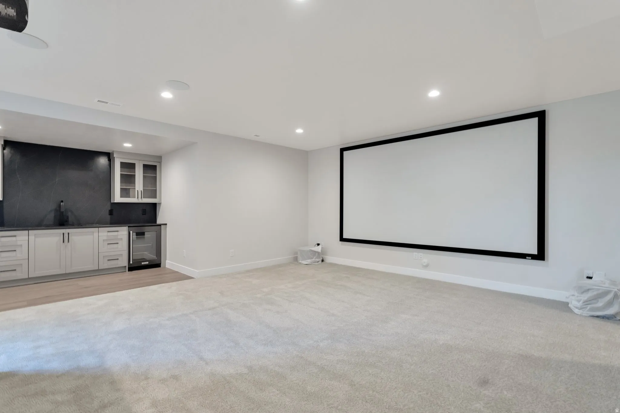 Spacious entertainment room featuring a large projection screen, recessed lighting, and a built-in wet bar with dark countertops, light cabinetry, and a beverage refrigerator