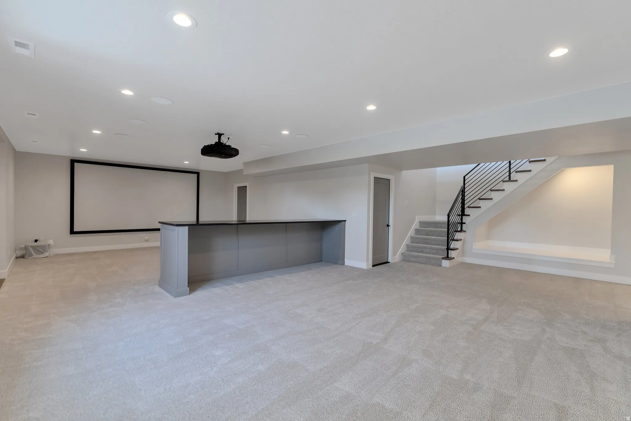 Expansive lower level featuring a built-in projector and large screen, a bar area with dark countertop, light grey carpeting, recessed lighting, and an open staircase with black metal railings