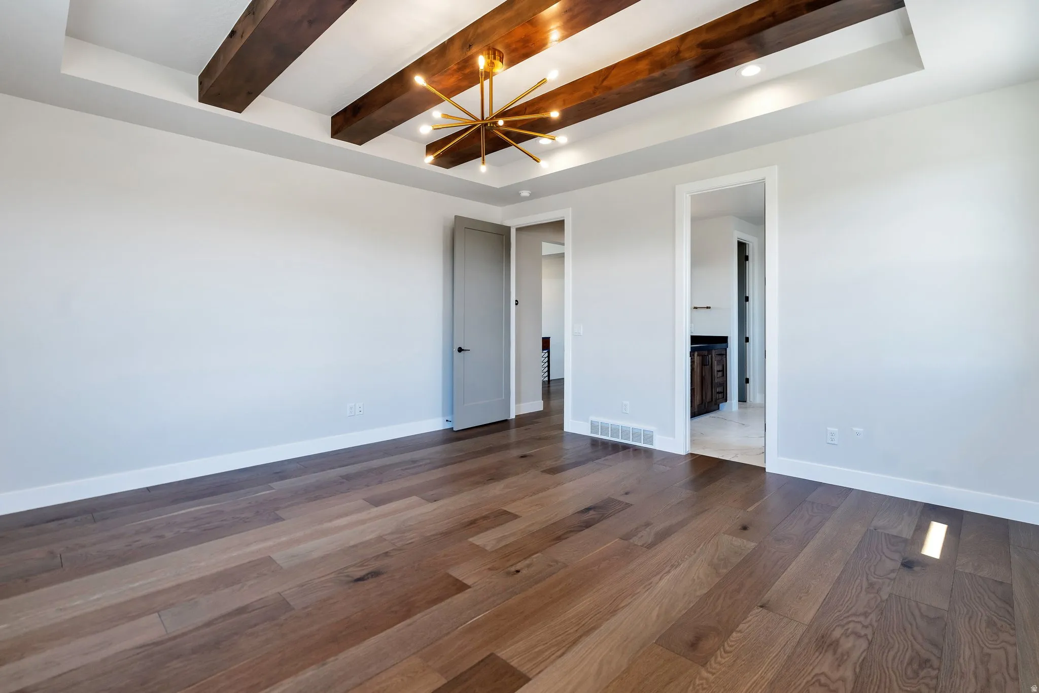Spacious room featuring wood-finish flooring, a coffered ceiling with dark wood beams, recessed lighting, and a contemporary brass chandelier