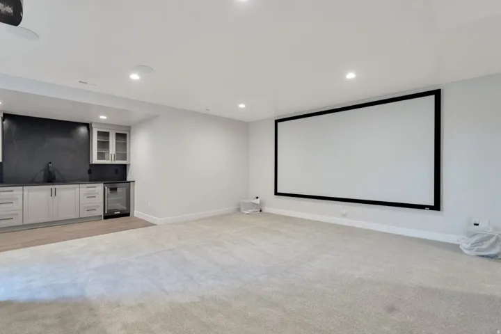 Spacious entertainment room featuring a large projection screen, recessed lighting, and a built-in wet bar with dark countertops, light cabinetry, and a beverage refrigerator