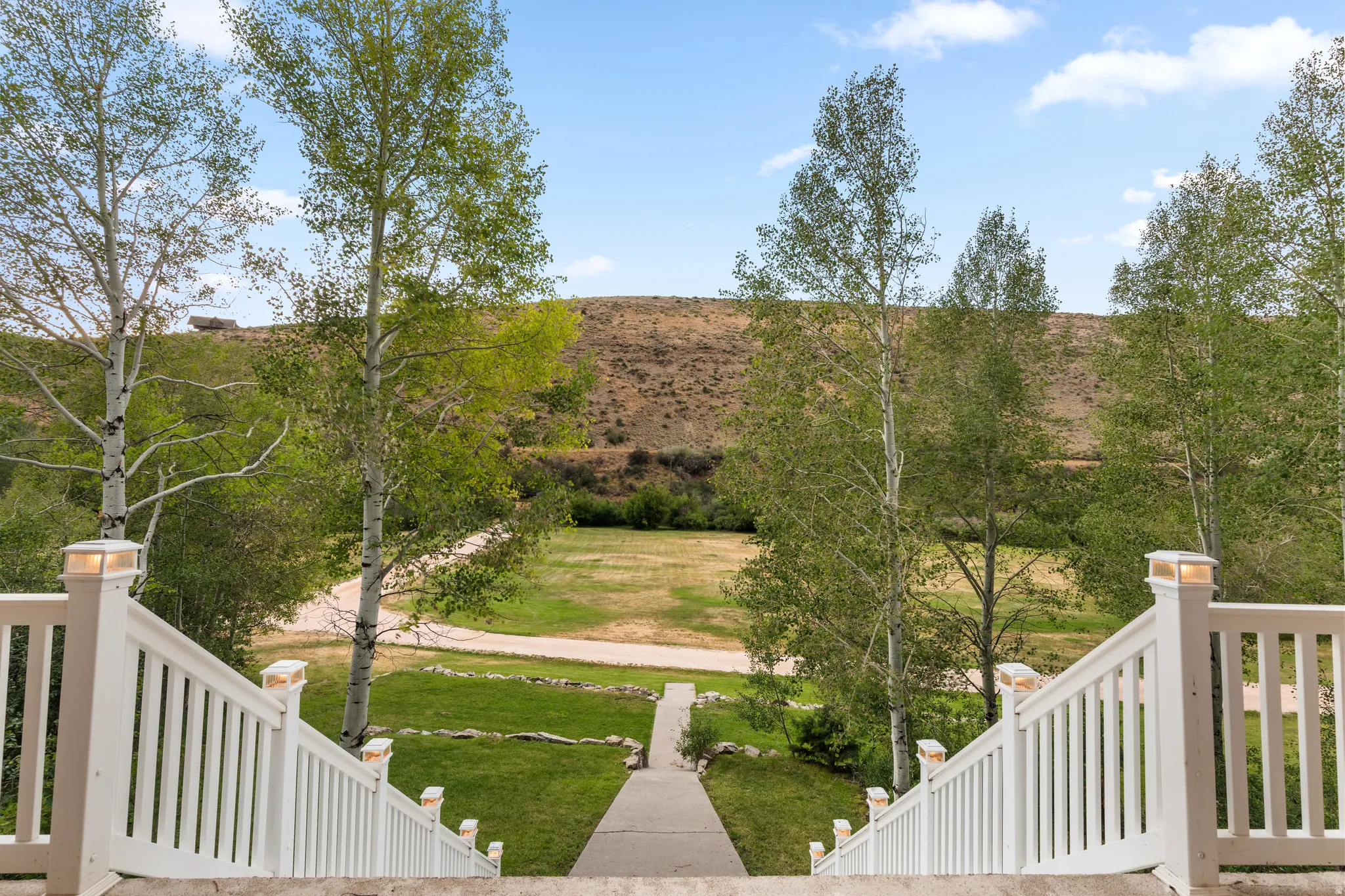 Outdoor staircase with white railings and post cap lights, leading to a large grass area with mature trees and a gravel path