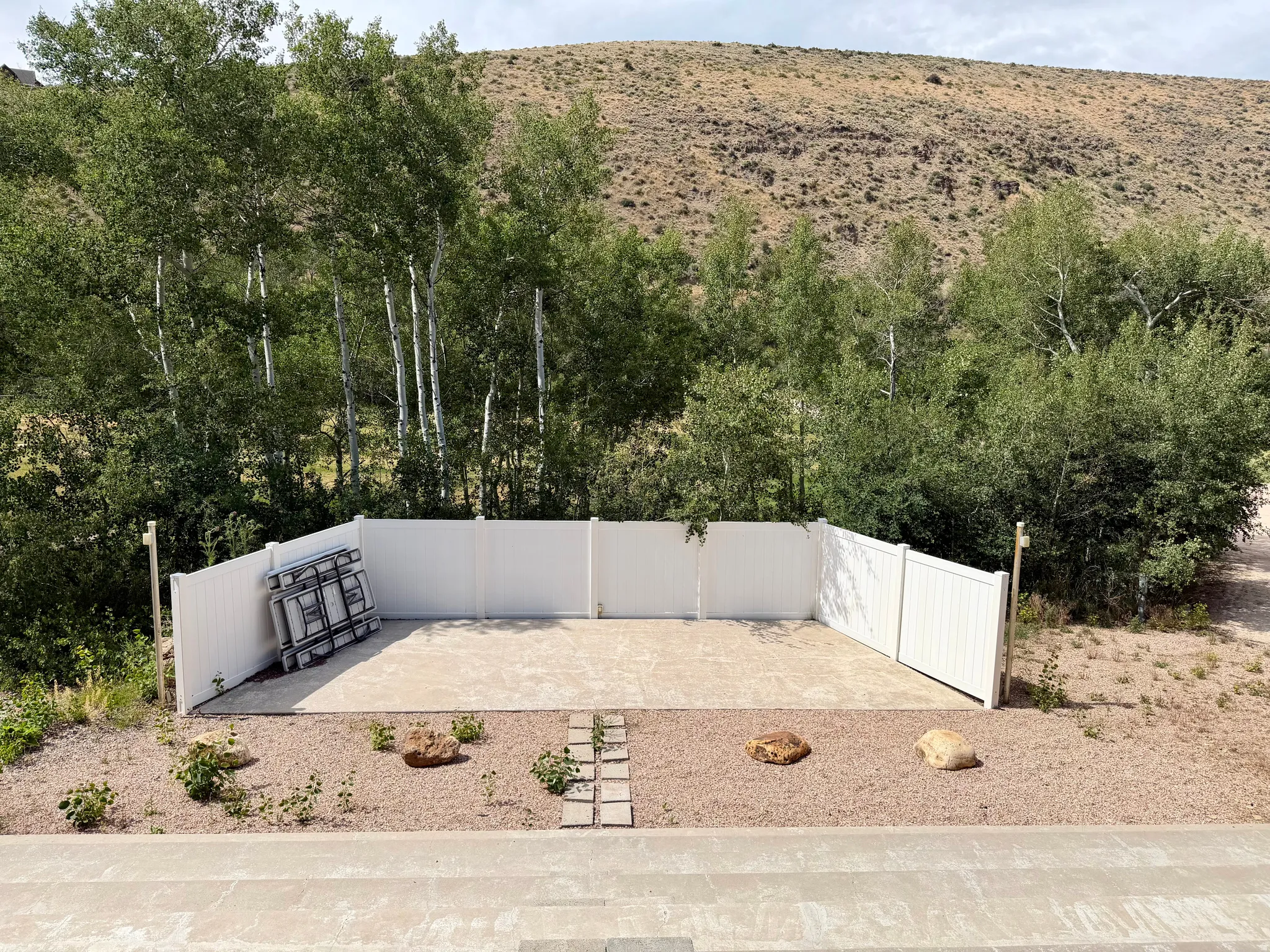 Outdoor paved amphitheater area featuring a white privacy fence, gravel landscaping with decorative rocks, and mature trees in the background