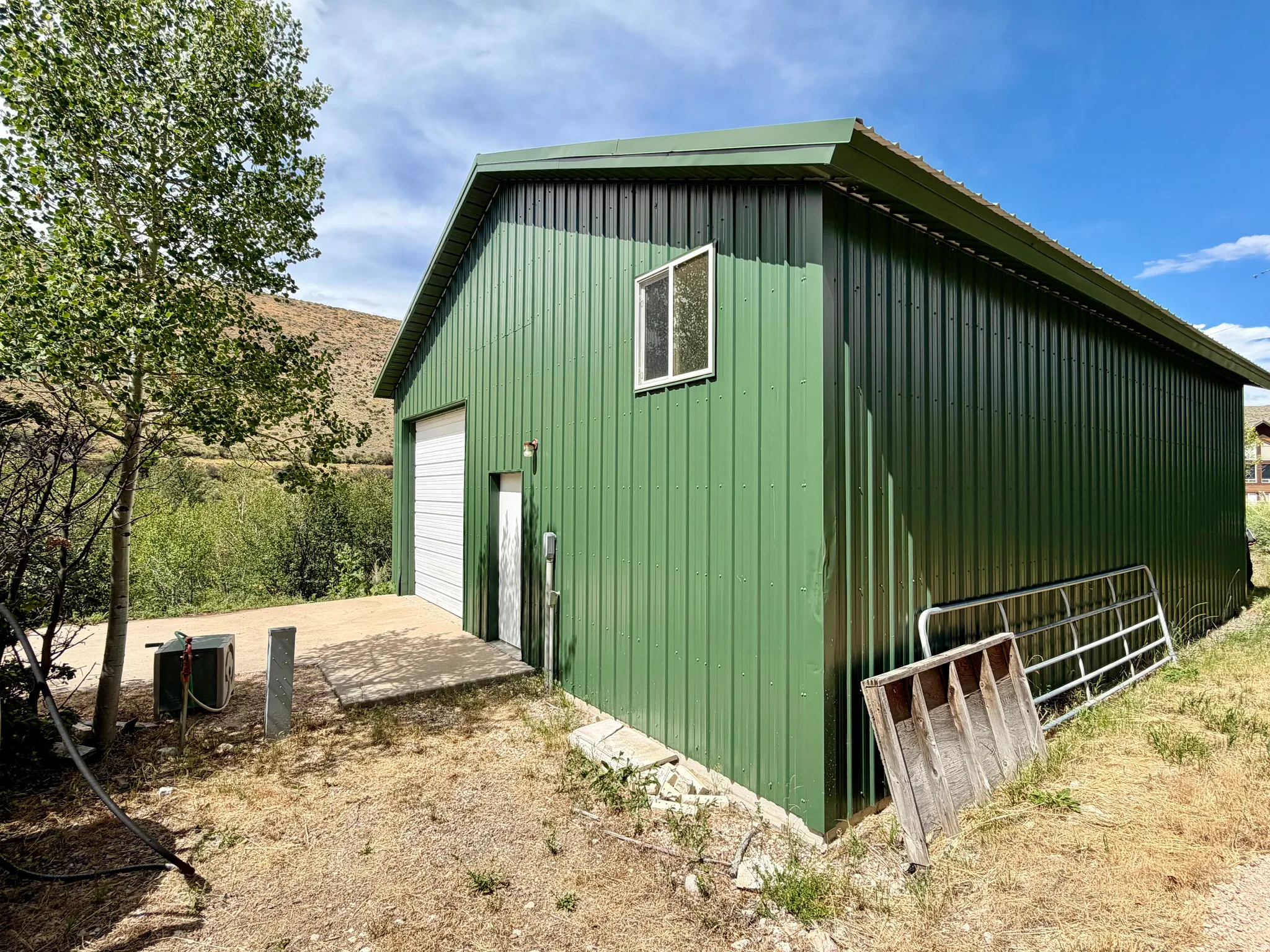 Green corrugated metal pavilion featuring a white roll-up garage door, a single window, and a concrete pad