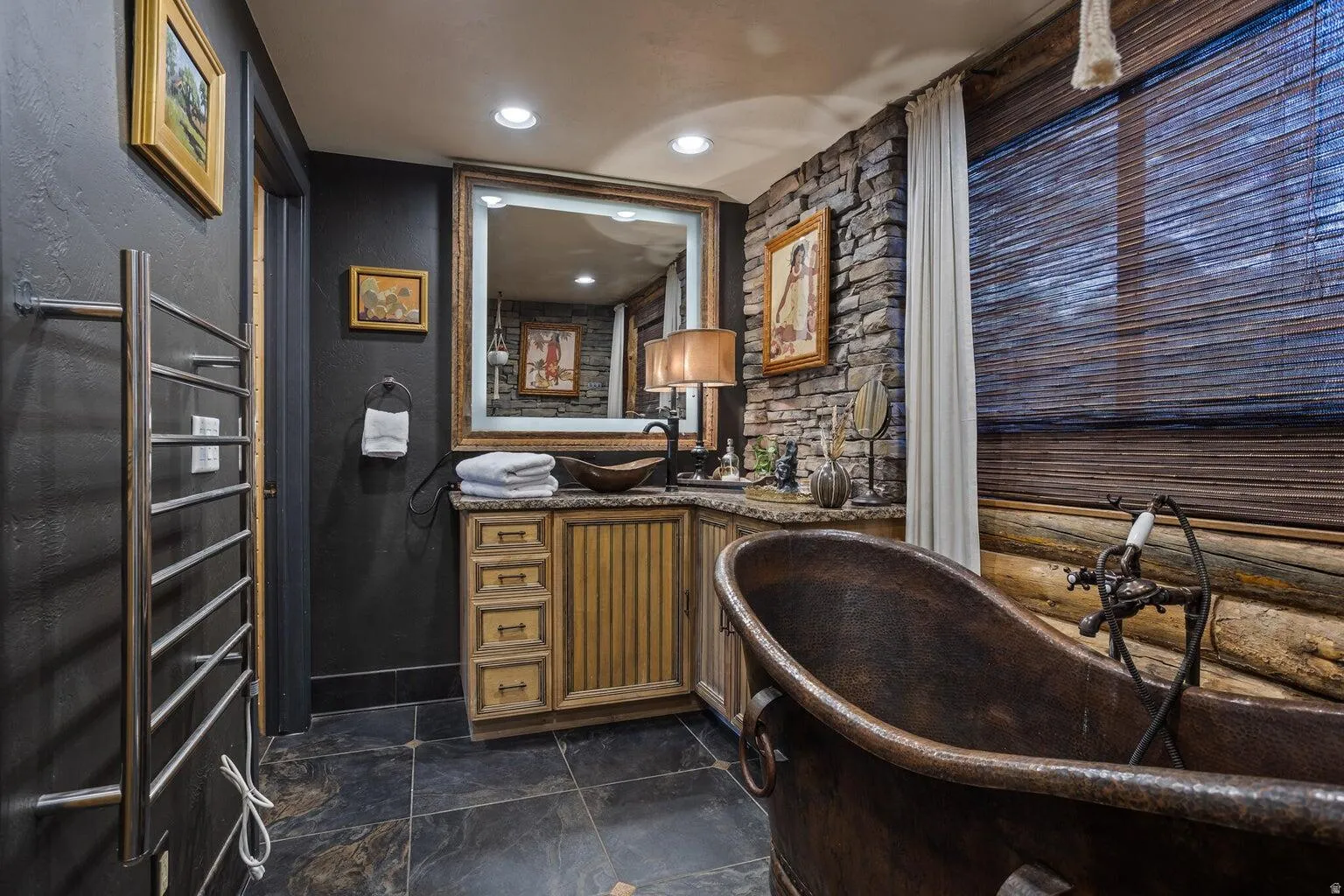Bathroom featuring a freestanding bronze bathtub, stone accent wall, wood-finish vanity with vessel sink, and slate-finish flooring