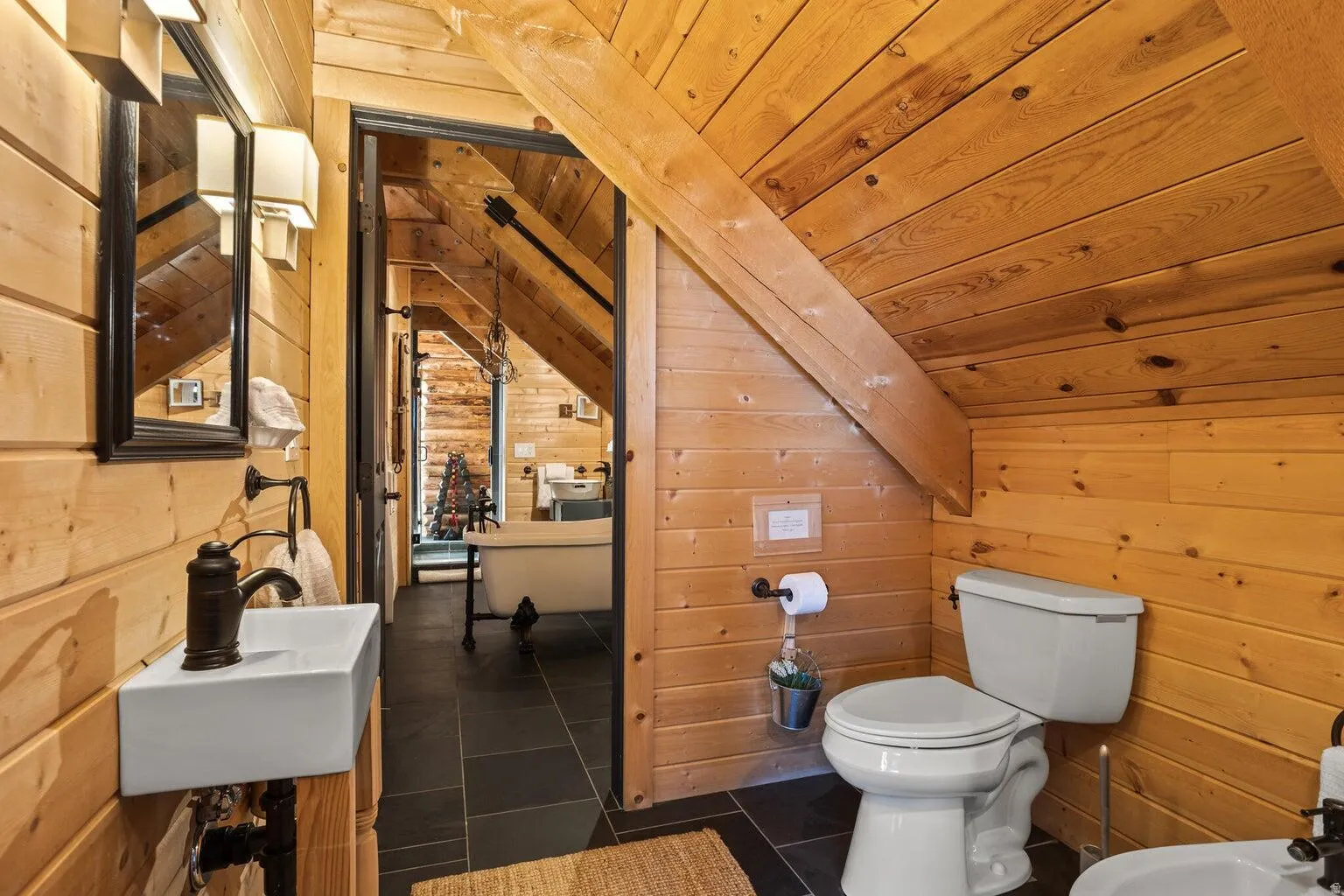 Bathroom with natural wood paneling, dark tile flooring, a wall-mounted rectangular sink, and a bronze-finish faucet