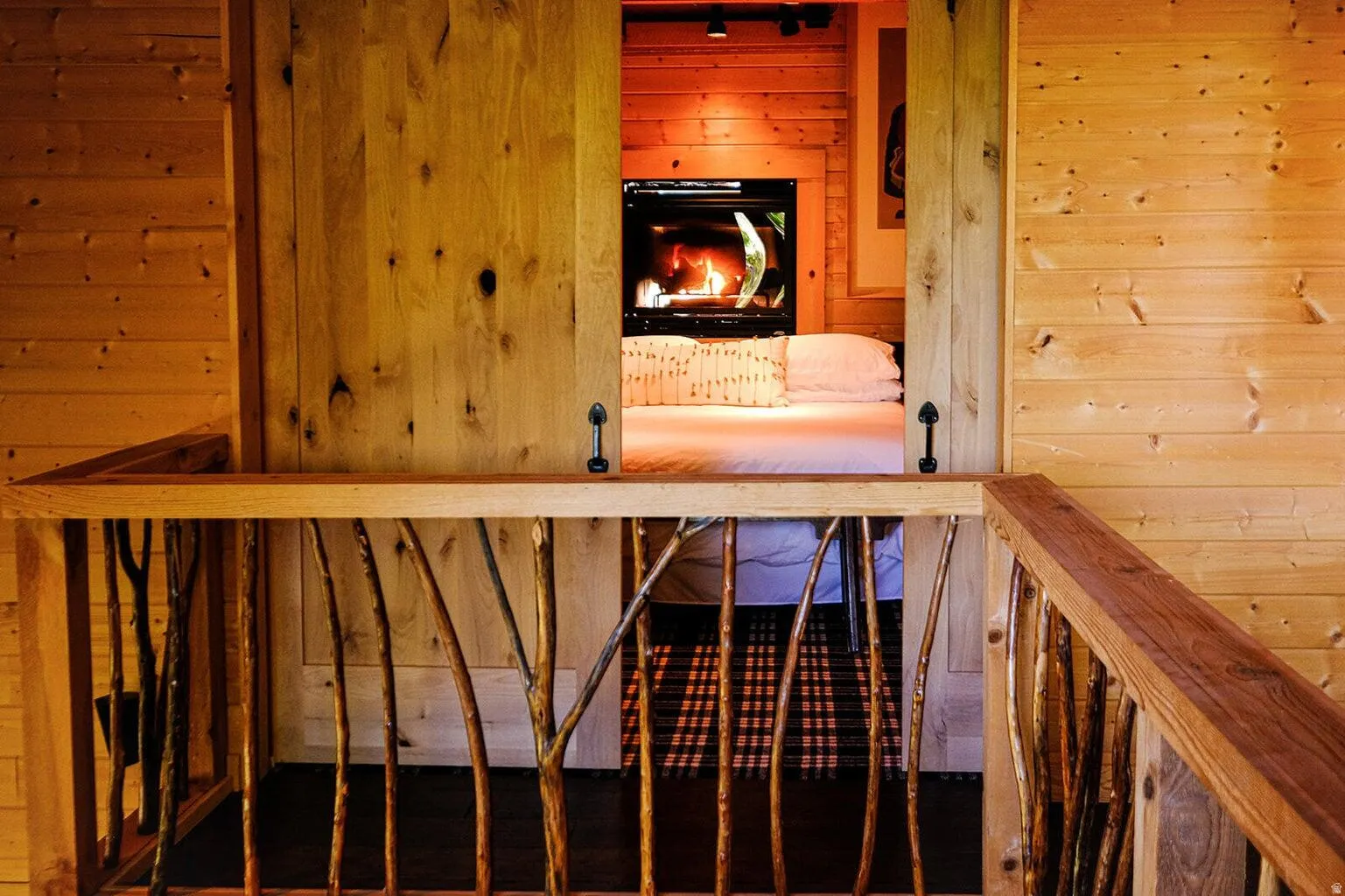 Natural wood railing with branch-style balusters opens to a room with a modern fireplace, wood-paneled walls, and track lighting