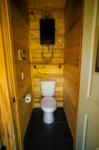 Compact lavatory featuring horizontal wood paneling, black floor tiling, a white toilet, and a wall-mounted water heater