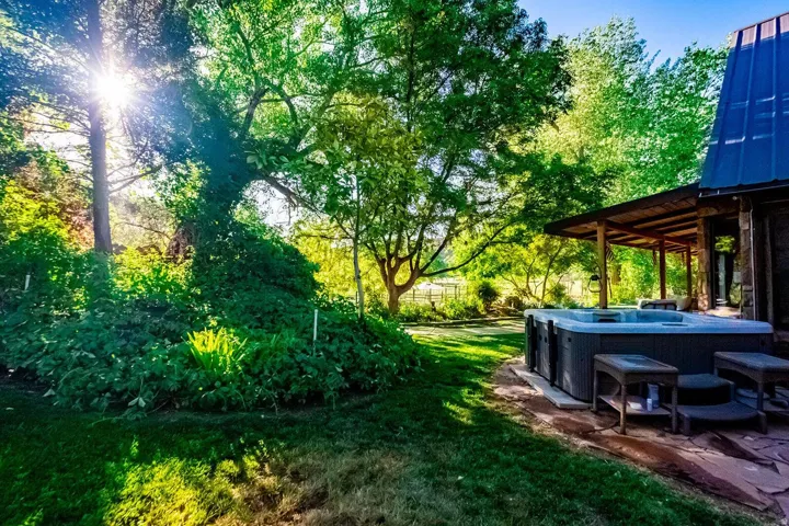 Expansive outdoor area featuring a hot tub on a flagstone patio, a covered porch with exposed beams, mature trees providing natural shade, and lush green landscaping