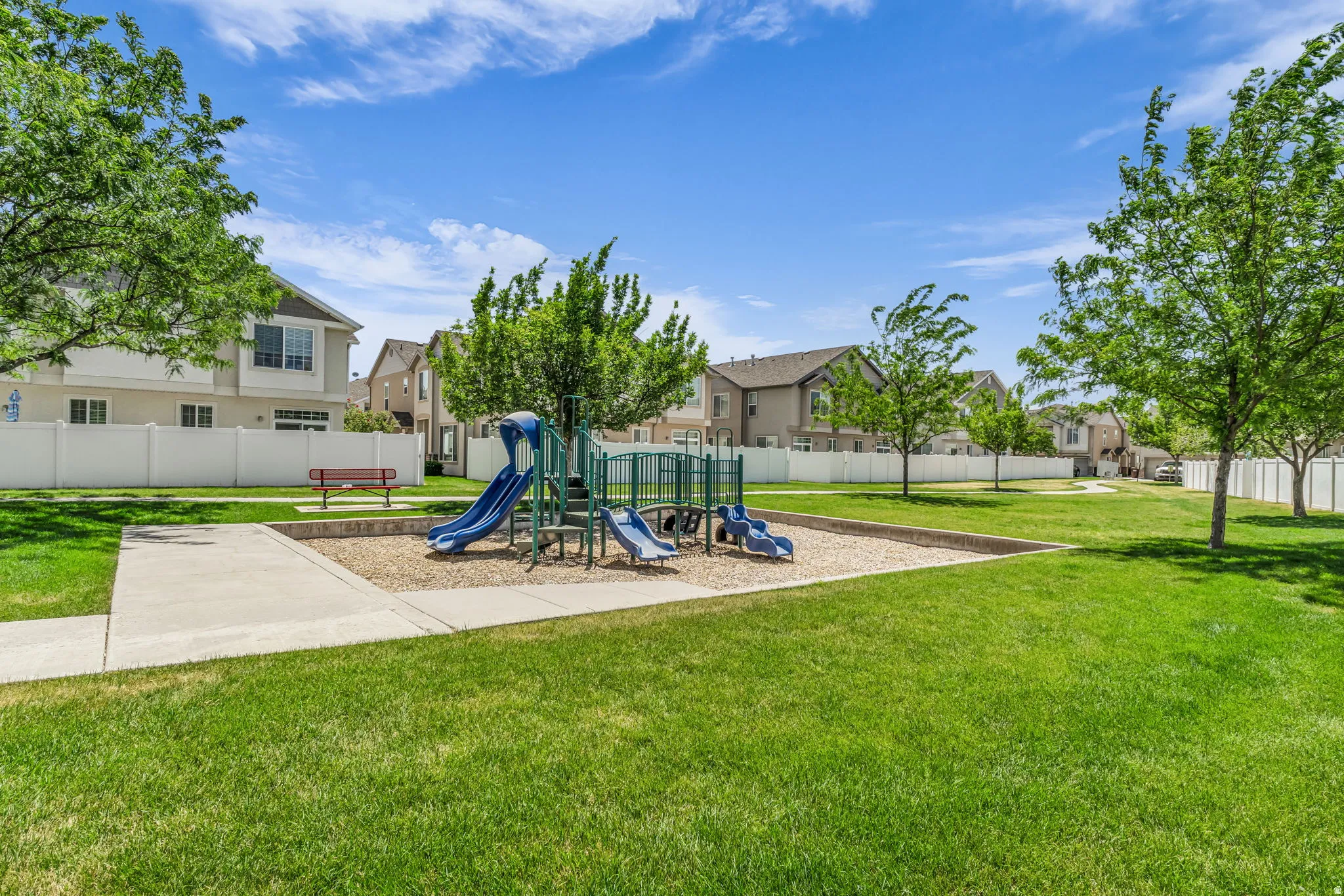 Open green space featuring a playground with blue slides and a climbing structure, surrounded by a white privacy fence