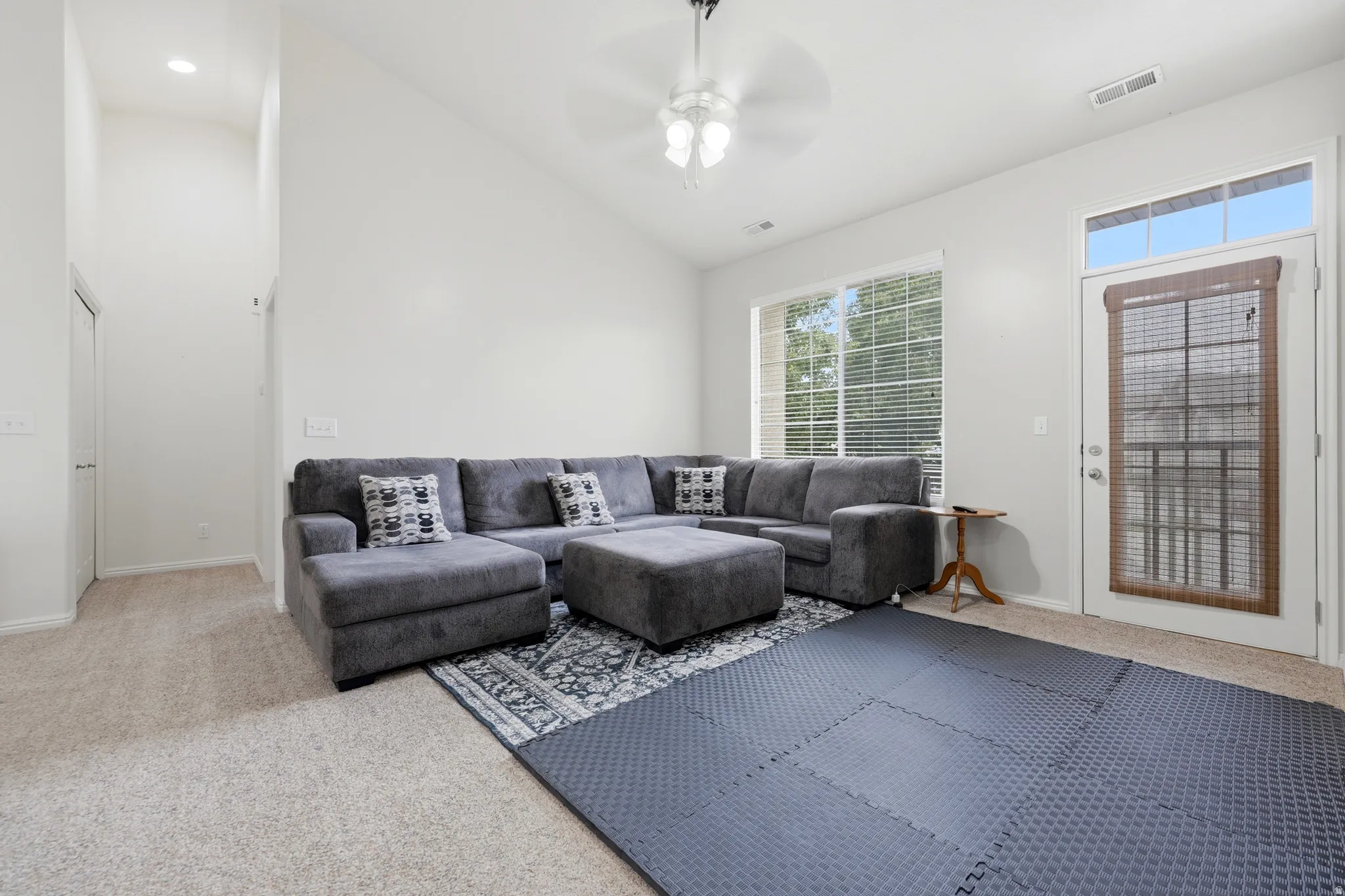 Open living space featuring a vaulted ceiling with a ceiling fan, a large window with blinds, and a glass-paneled door