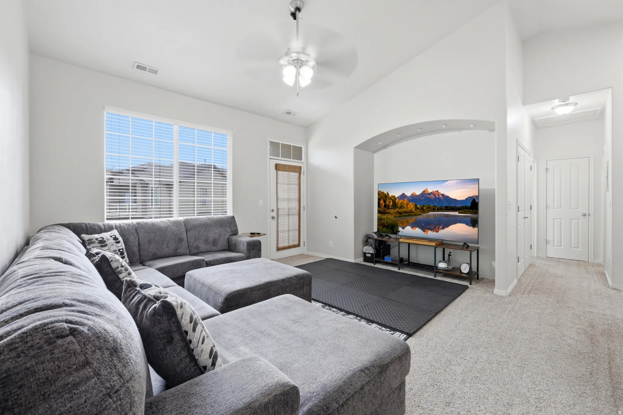 Open-concept living space featuring high vaulted ceilings, large windows with blinds, a ceiling fan, an arched alcove, and light-toned carpeting throughout