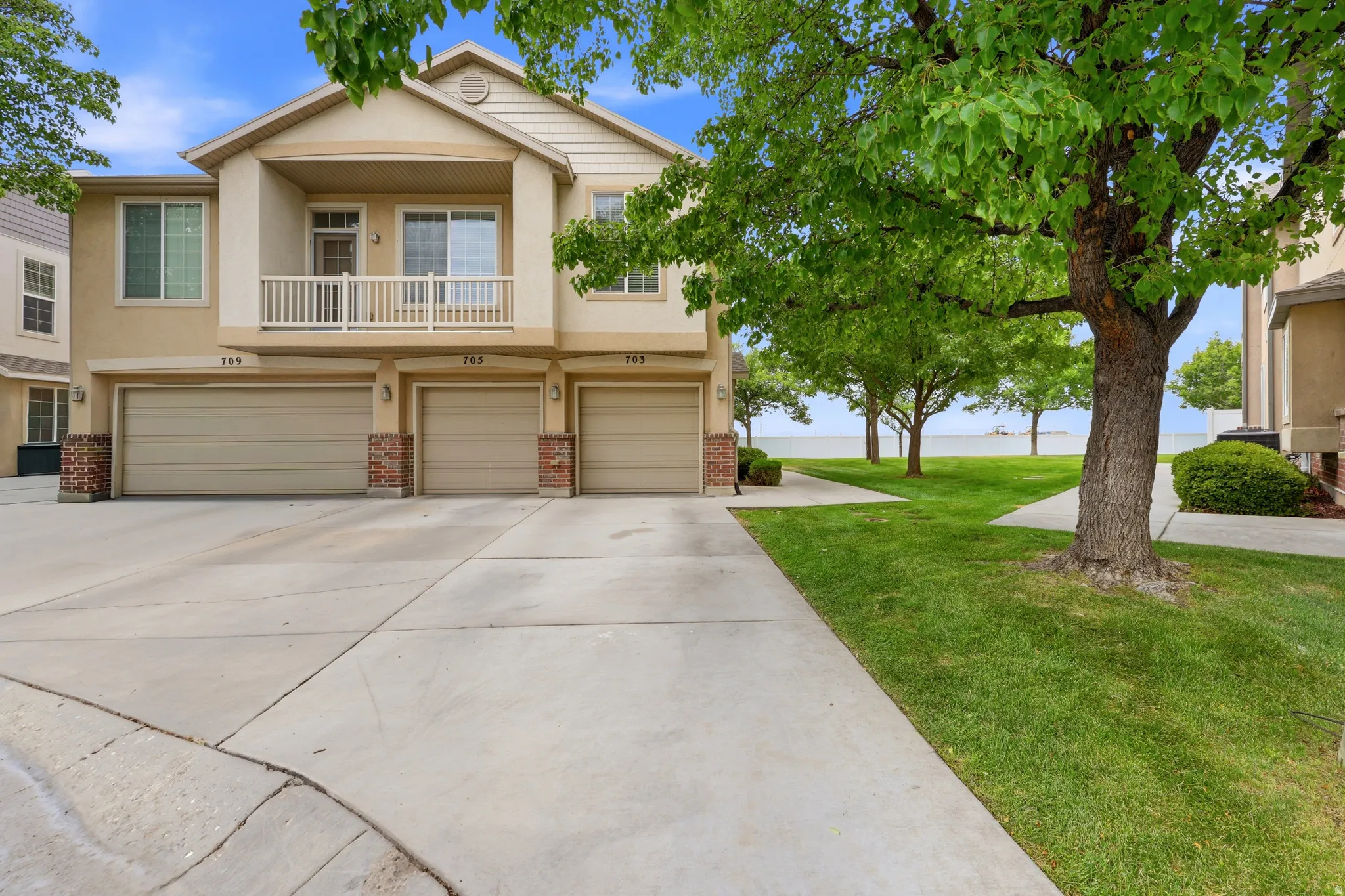 Townhome exterior featuring a stucco finish, a second-story balcony with white railing, individual garages with brick accents, concrete driveways, and a grassy lawn with mature trees