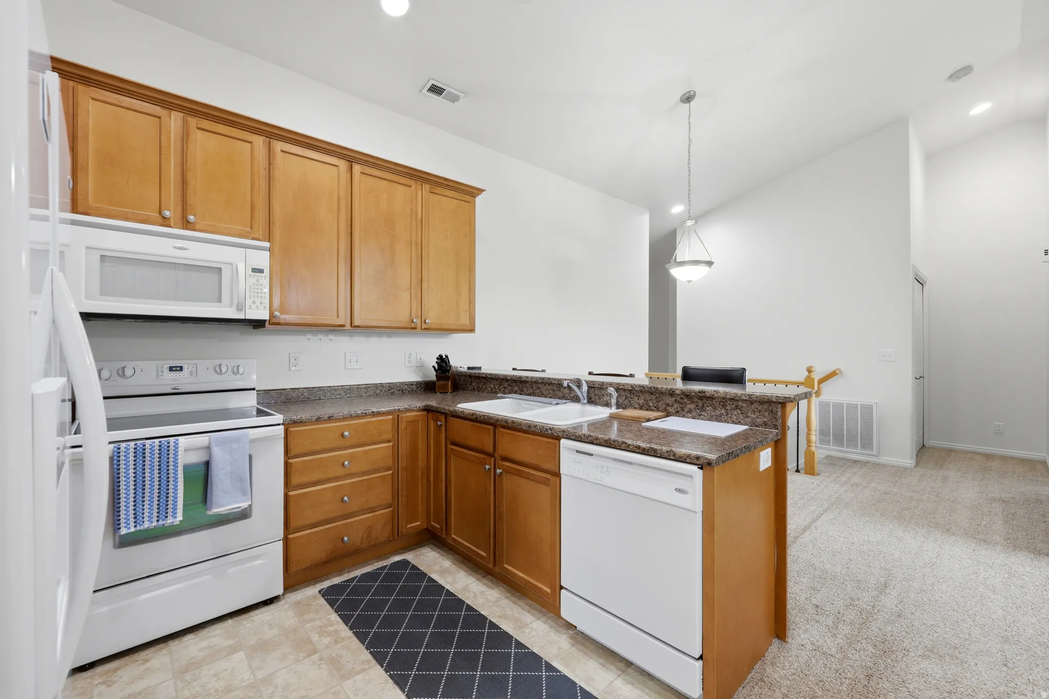 Well-appointed kitchen featuring light wood cabinetry, white appliances, a double basin sink, stone-finish countertops, and a breakfast bar