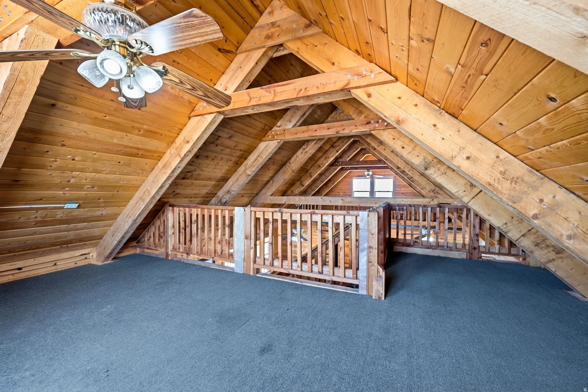 Loft space featuring exposed wood beams, wood paneling, dark carpet, a ceiling fan with integrated lighting, and a window providing natural illumination