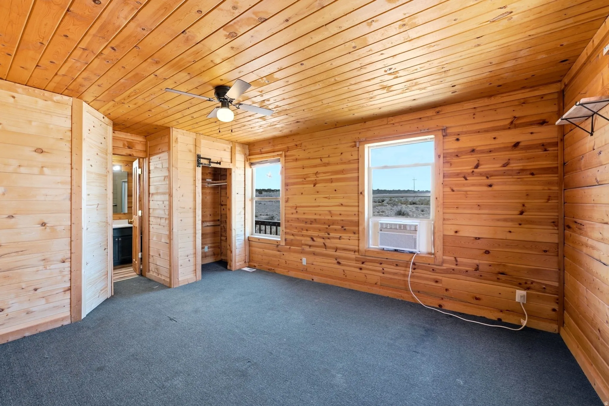 Wood-paneled room featuring a ceiling fan, two windows, carpet flooring, a built-in closet with a sliding barn door, and an entryway to an adjoining space with a vanity