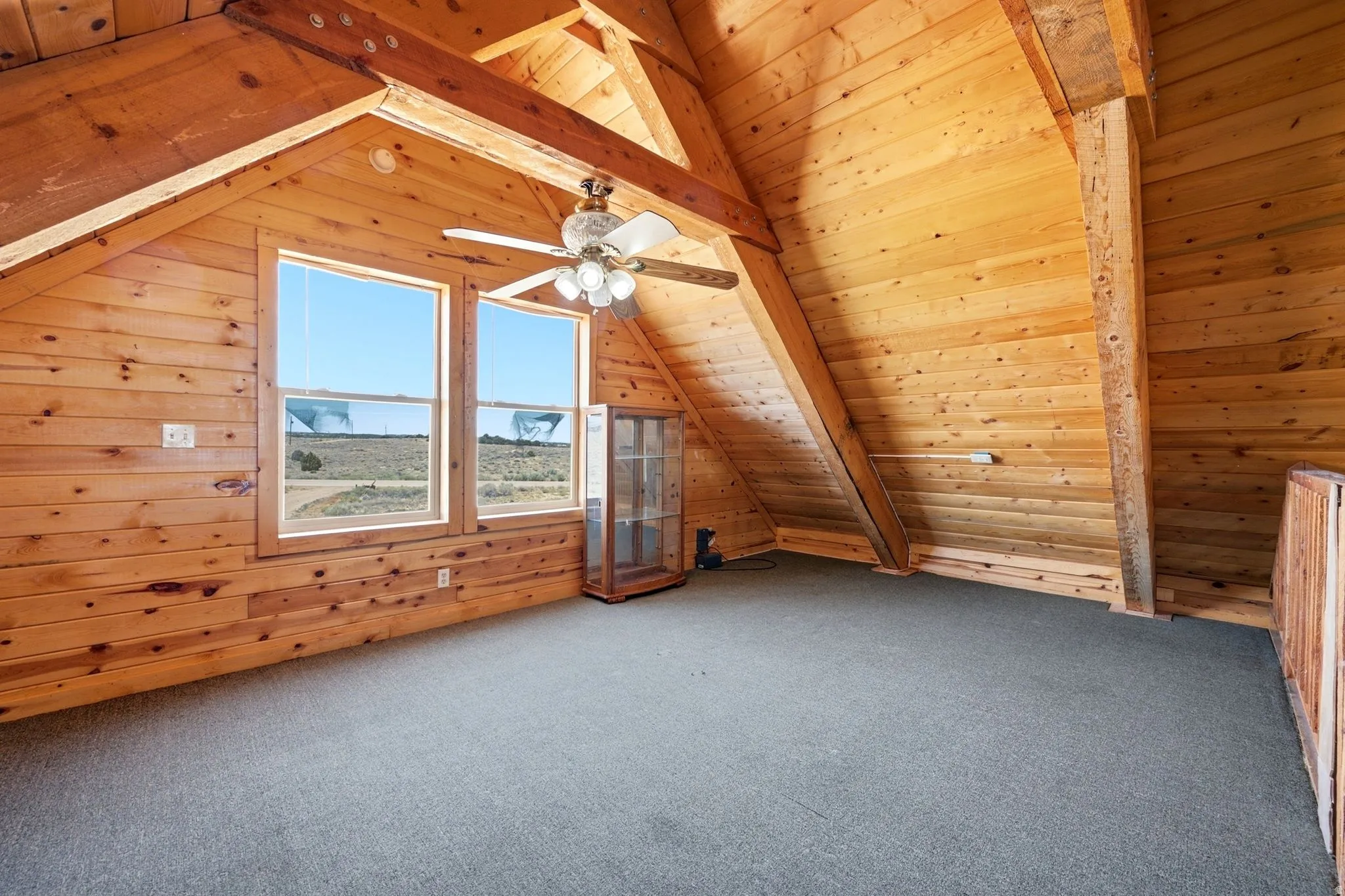 Natural wood-paneled walls and ceilings featuring exposed wood beams, two large windows, a ceiling fan with integrated lighting, and neutral carpeting