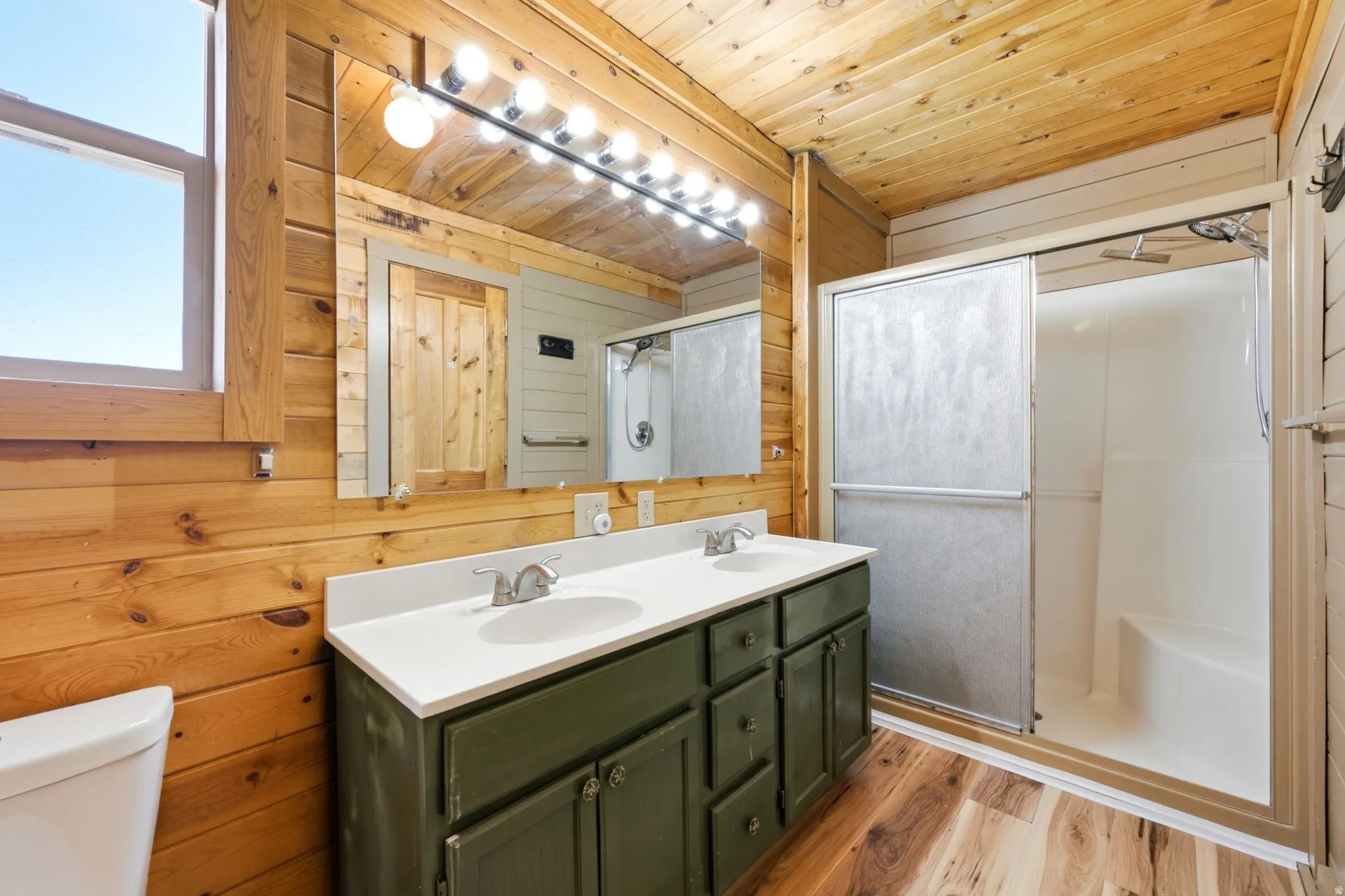 Bathroom featuring a double vanity with a white countertop and green cabinetry, a shower with sliding doors and an integrated bench, wood-finish flooring, and wood paneling on the walls and ceiling