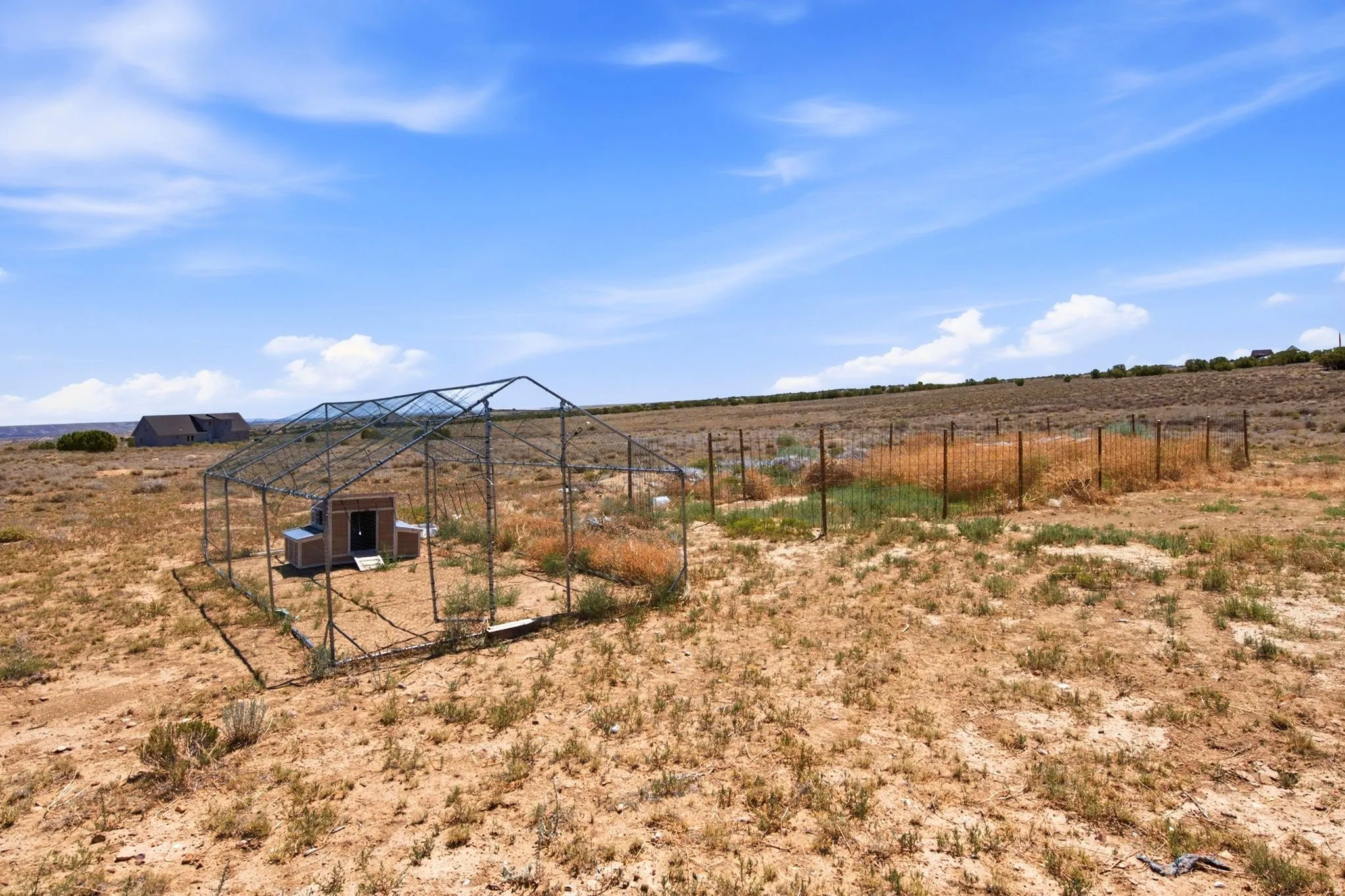 Expansive dryland property featuring a fenced enclosure with a small outbuilding, additional partial fencing, and distant structures under a clear sky
