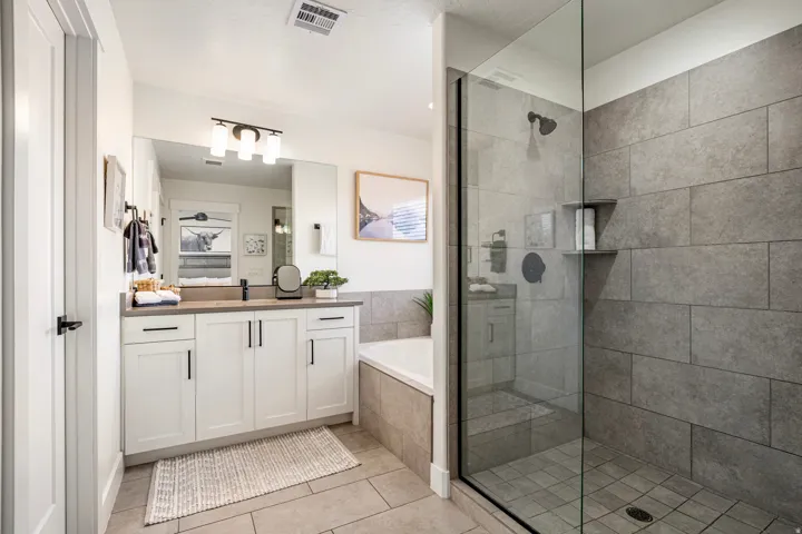 Spacious bathroom featuring a glass-enclosed shower with gray tile surround, a built-in bathtub, and a double vanity with white cabinetry and dark countertops