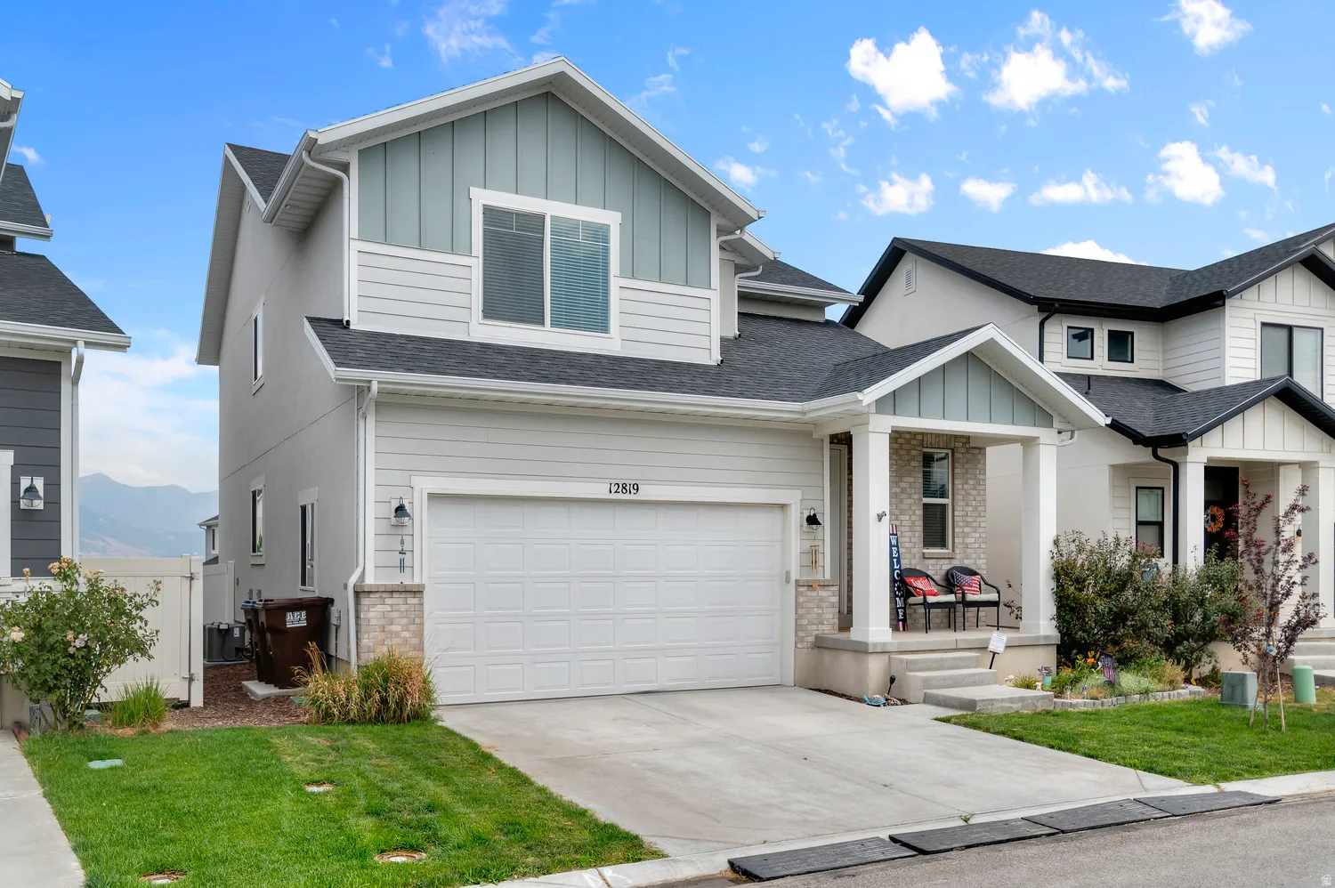 Contemporary two-story residence featuring a two-car garage, a covered front porch with stone accent pillars, and a light-toned exterior with board and batten siding details