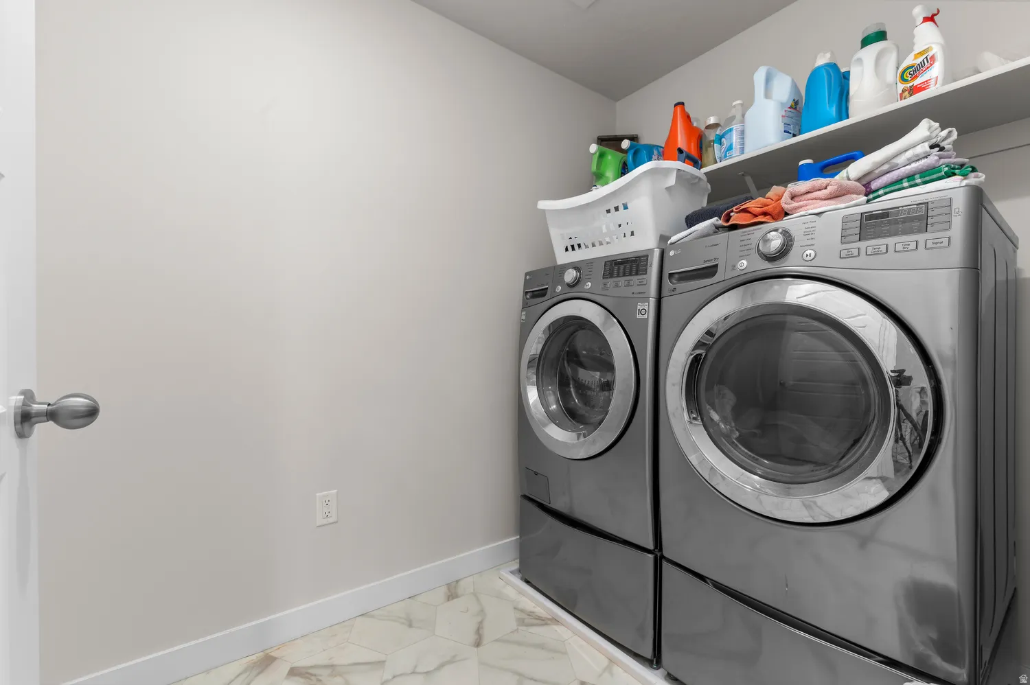 Dedicated laundry area featuring front-loading washer and dryer units, a wall-mounted storage shelf, and light-toned tile flooring